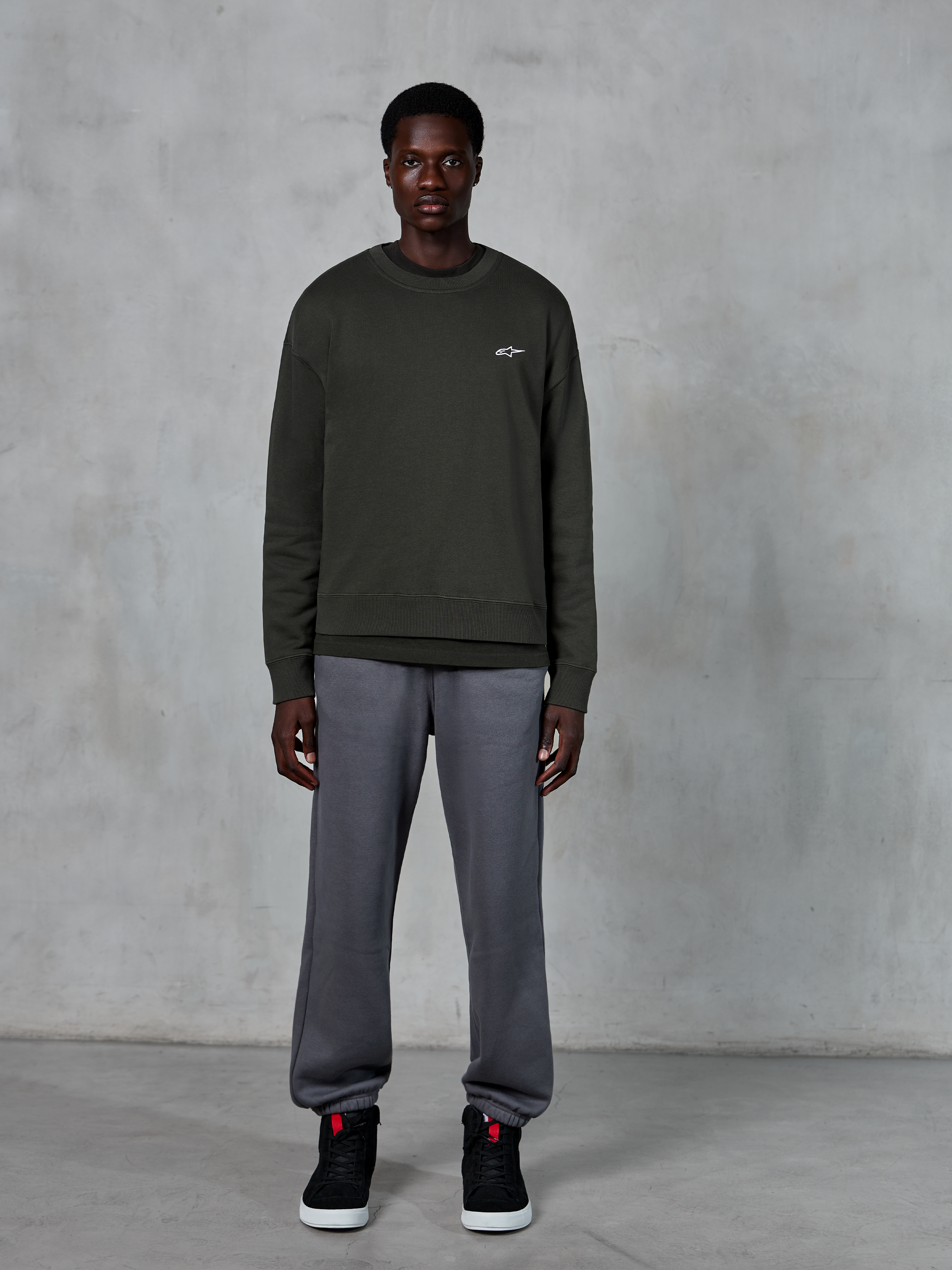 Alpinestars Essentials Ageless Crew, dunkelgrünes Rundhals-Sweatshirt mit kleinem weißem Logo auf der Brust, getragen von einem Model mit grauer Jogginghose und schwarzen High-Top-Sneakern, lässige Lifestyle-Bekleidung