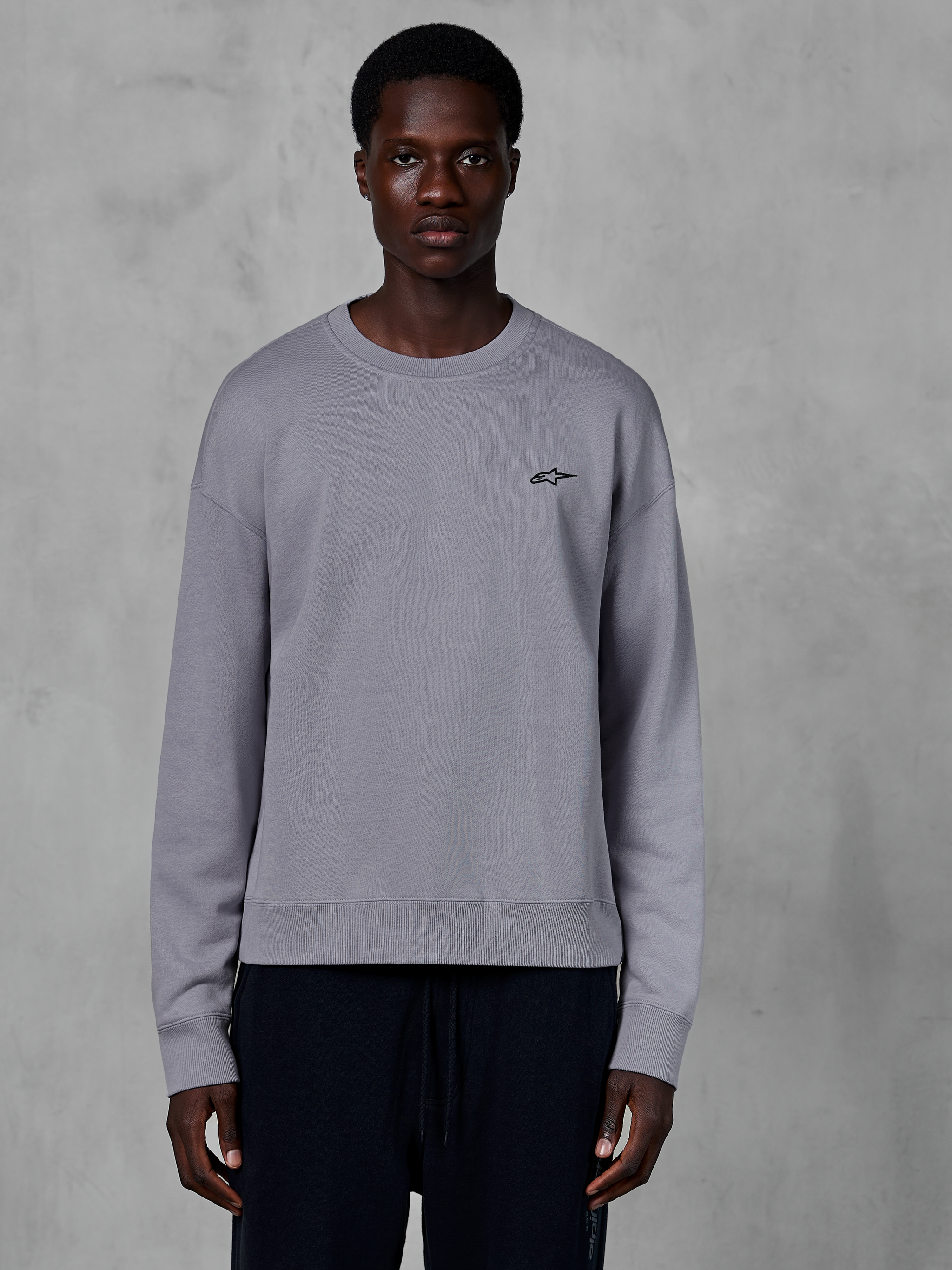 Alpinestars Essentials Ageless Crew, sweat-shirt à col rond argenté porté par un mannequin, avec un petit logo étoile noir sur la poitrine, coupe décontractée avec poignets et ourlet côtelés, style streetwear décontracté inspiré du sport automobile