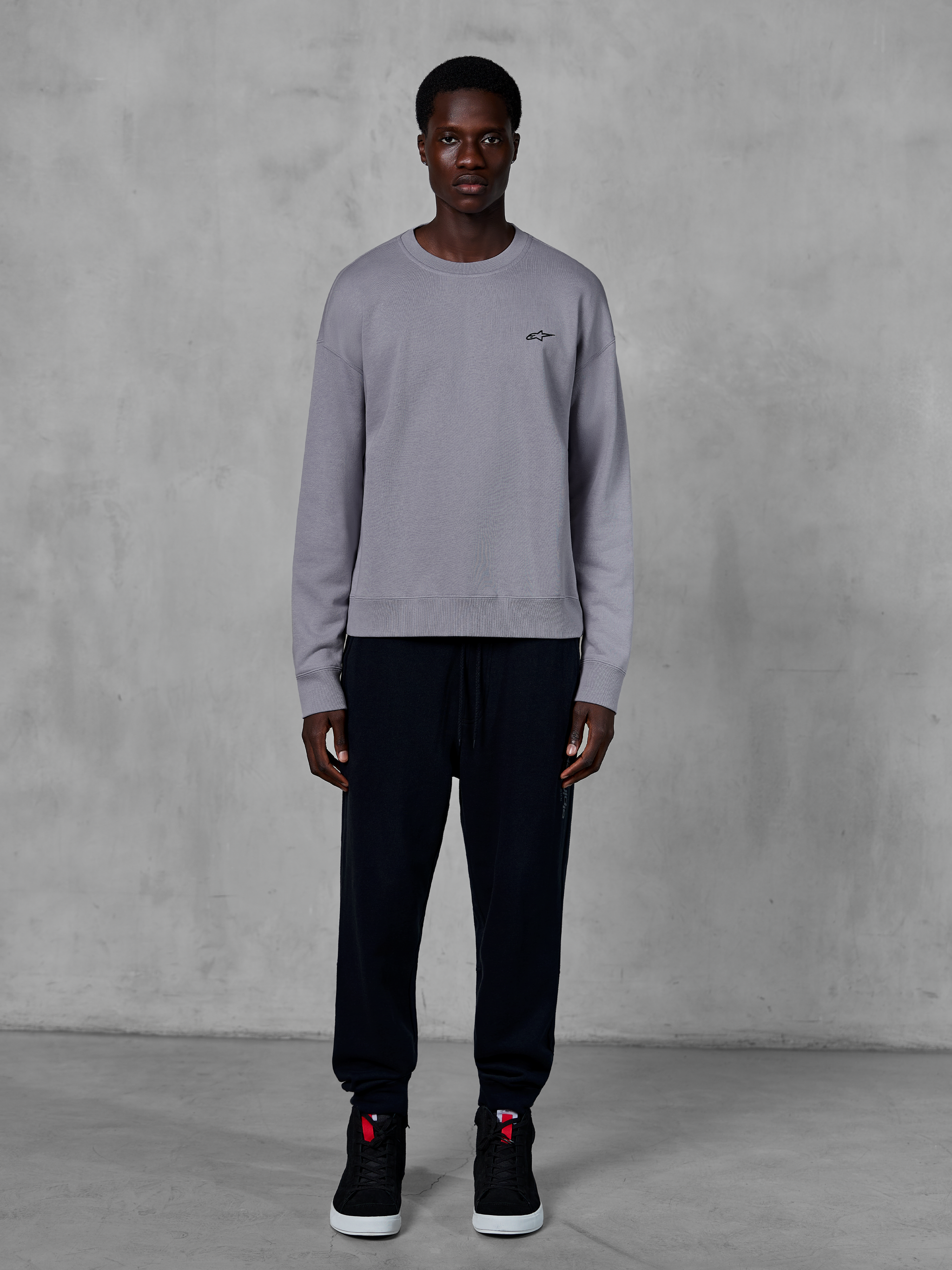 Alpinestars Essentials Ageless Crew, sweat-shirt à col rond argenté, porté par un mannequin avec un jogging noir et des baskets noires, arborant un petit logo Alpinestars noir sur la poitrine, streetwear décontracté inspiré du sport automobile