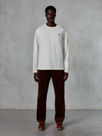 Alpinestars Essentials Ageless LS Tee, t-shirt à manches longues, blanc cassé, porté par un mannequin avec un pantalon en velours côtelé marron et des bottes en cuir marron, avec un petit logo noir sur la poitrine, coupe décontractée contemporaine, style streetwear décontracté