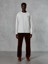 Alpinestars Essentials Ageless LS Tee, t-shirt à manches longues, blanc cassé, porté par un mannequin avec un pantalon en velours côtelé marron et des bottes en cuir marron, avec un petit logo noir sur la poitrine, coupe décontractée contemporaine, style streetwear décontracté