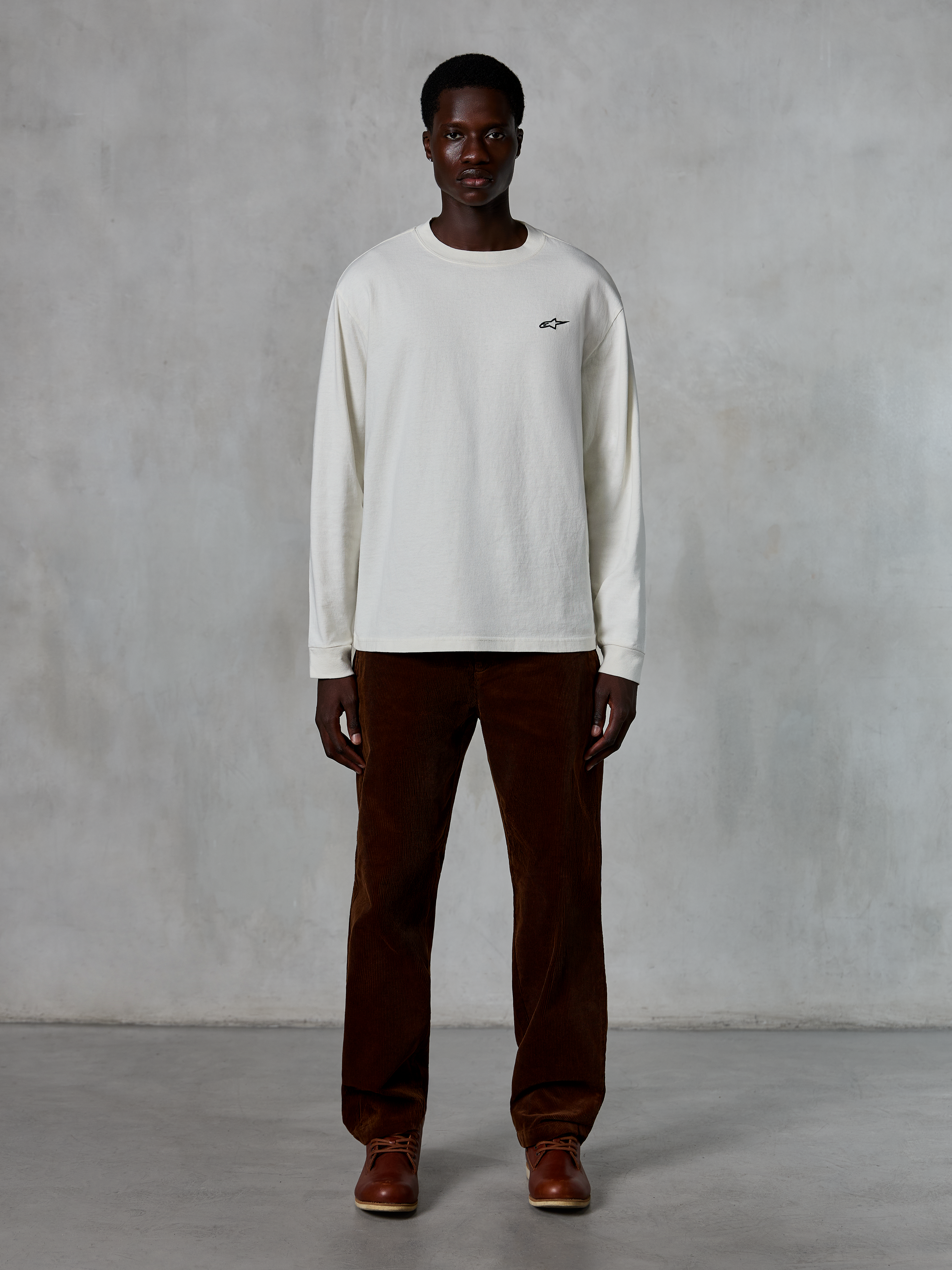 Alpinestars Essentials Ageless LS Tee, t-shirt à manches longues, blanc cassé, porté par un mannequin avec un pantalon en velours côtelé marron et des bottes en cuir marron, avec un petit logo noir sur la poitrine, coupe décontractée contemporaine, style streetwear décontracté