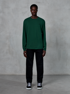 T-shirt à manches longues Alpinestars Essentials Ageless, vert sapin, doté d'un petit logo noir sur la poitrine, porté par un mannequin avec un pantalon noir et des baskets, style streetwear décontracté