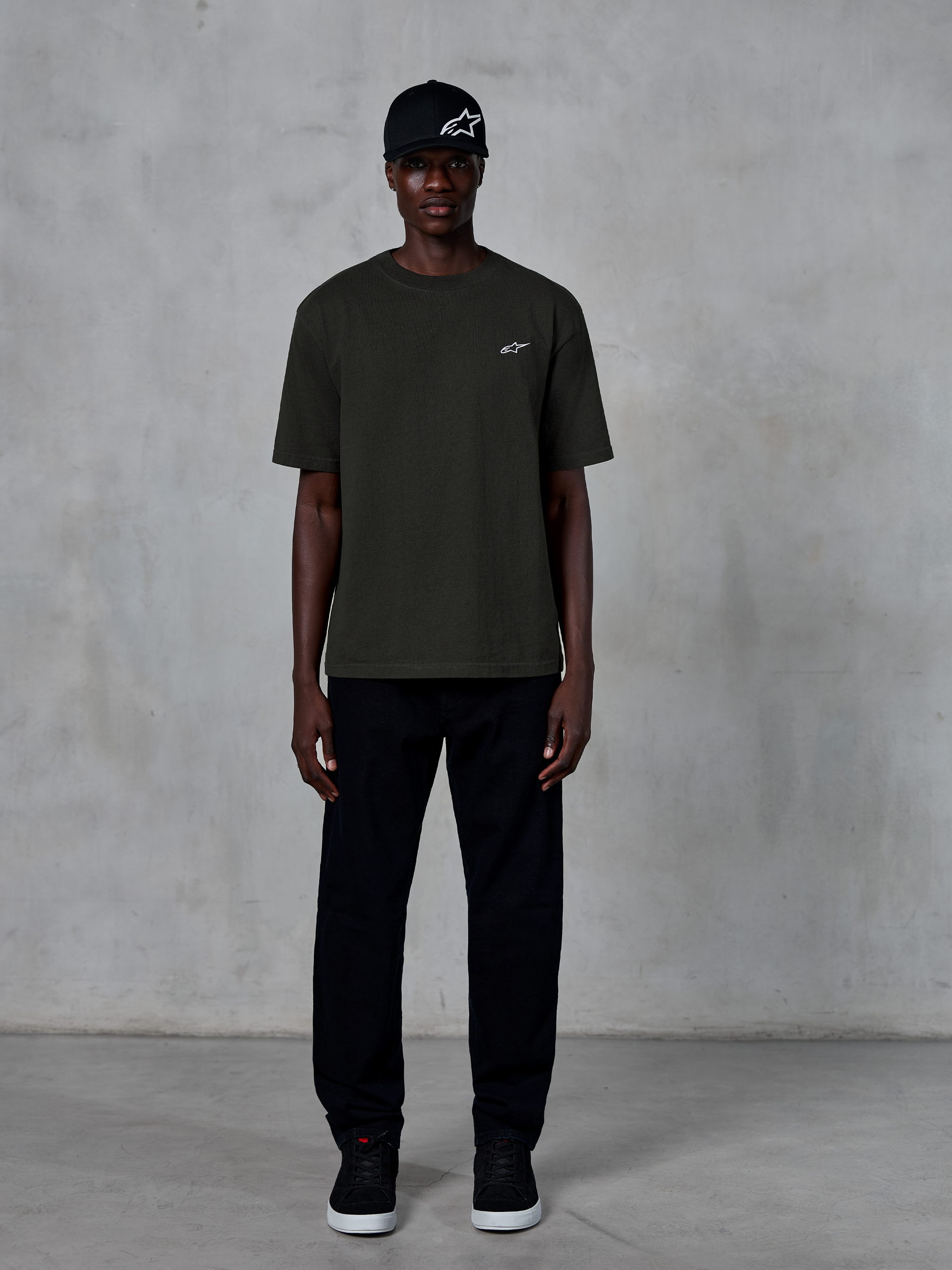 Alpinestars Essentials Ageless SS Tee, Kurzarm-T-Shirt, Off Black, mit einem kleinen weißen Alpinestars-Logo auf der Brust, getragen von einem Model mit schwarzer Kappe und Hose für einen cleanen Lifestyle-Look