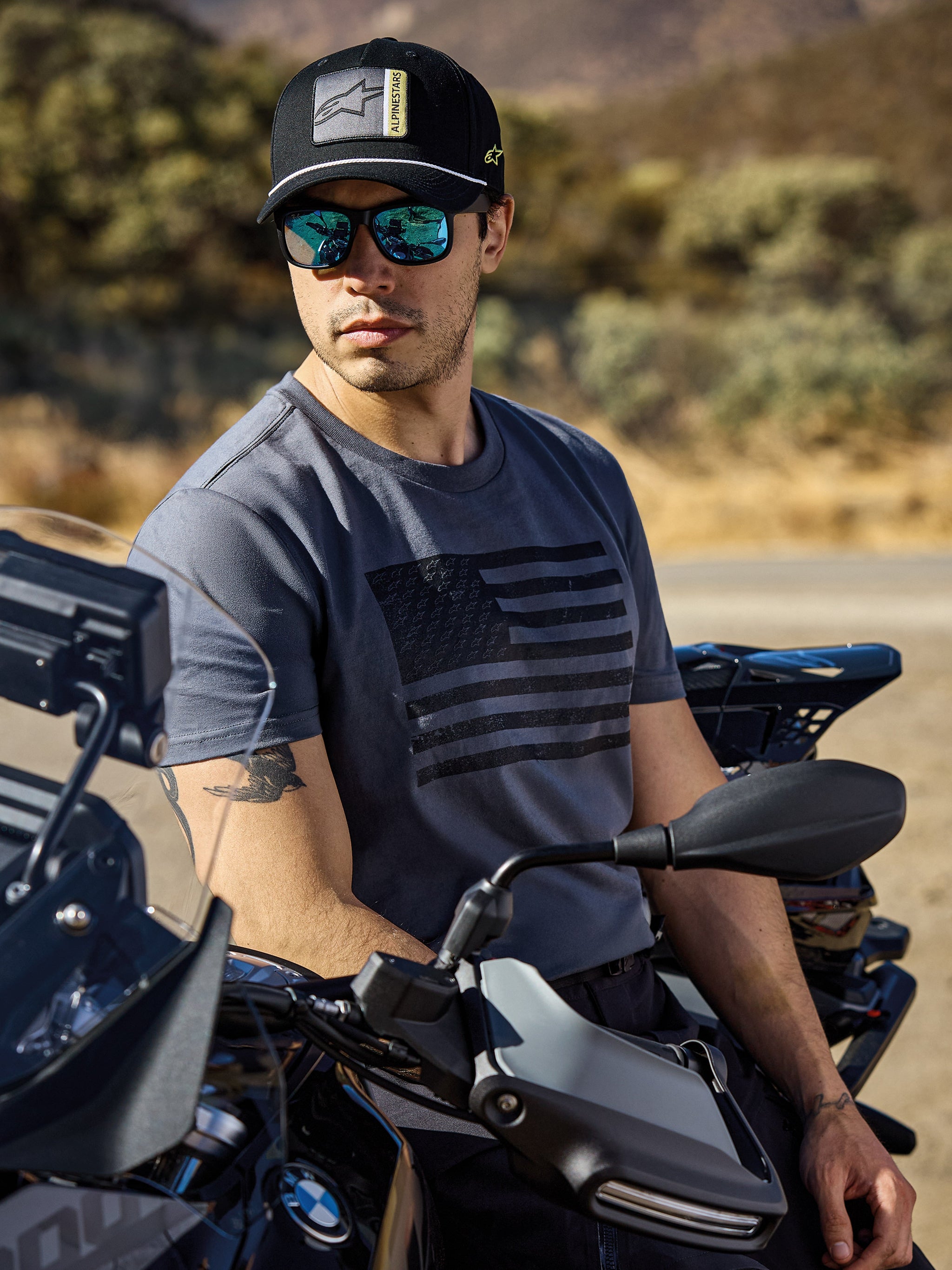 Alpinestars Waving CSF Tee, T-shirt met korte mouwen, charcoal, met een zwarte wapperende vlag-graphic op de borst, gedragen door een man op een motor in een zonnige buitenomgeving, casual motorsport lifestyle