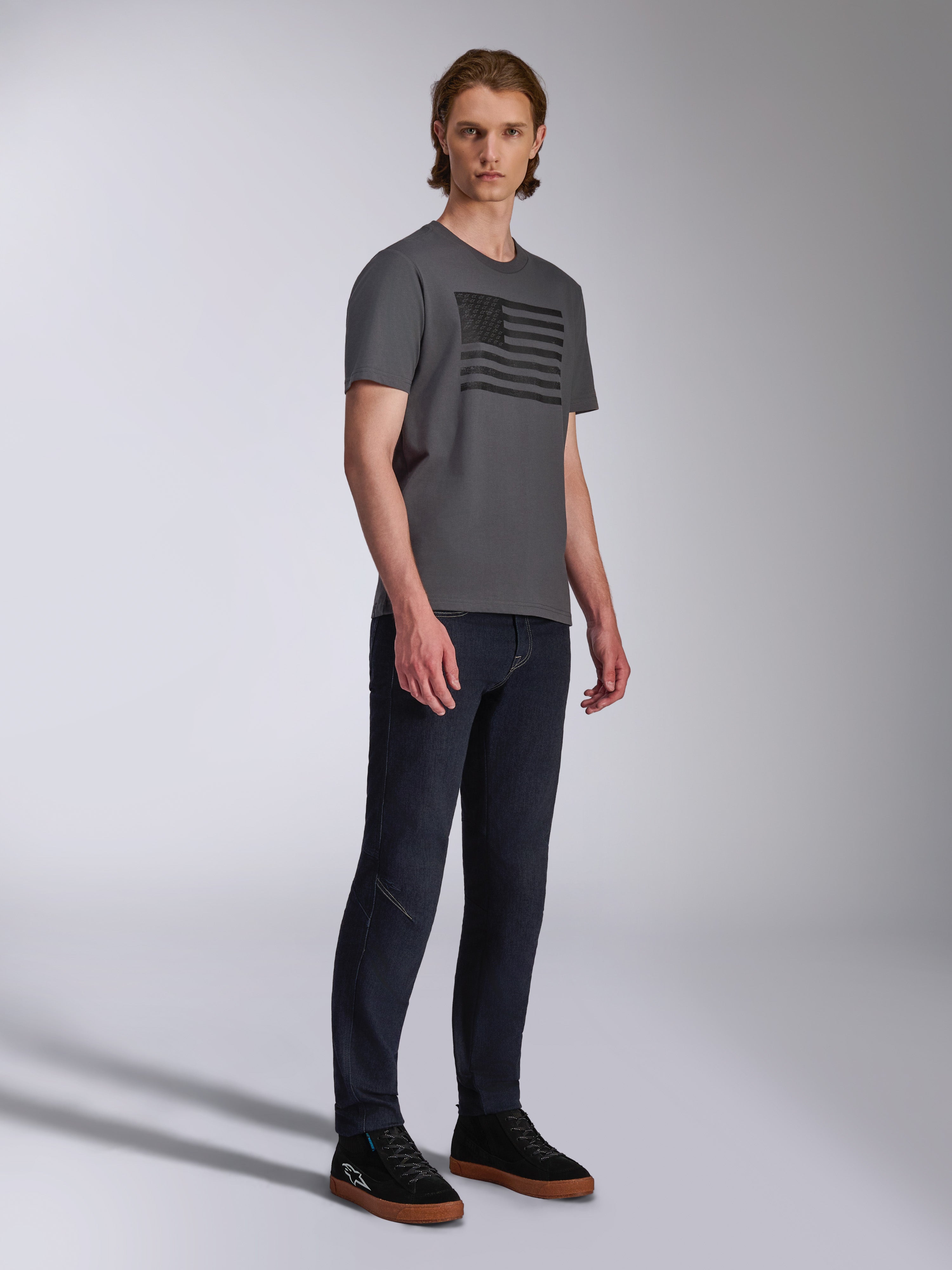 Alpinestars Waving CSF Tee, T-shirt met korte mouwen, antraciet, zwarte grafische print van een wapperende vlag, gedragen door een model met donkere jeans en sneakers, casual motorsport streetwear