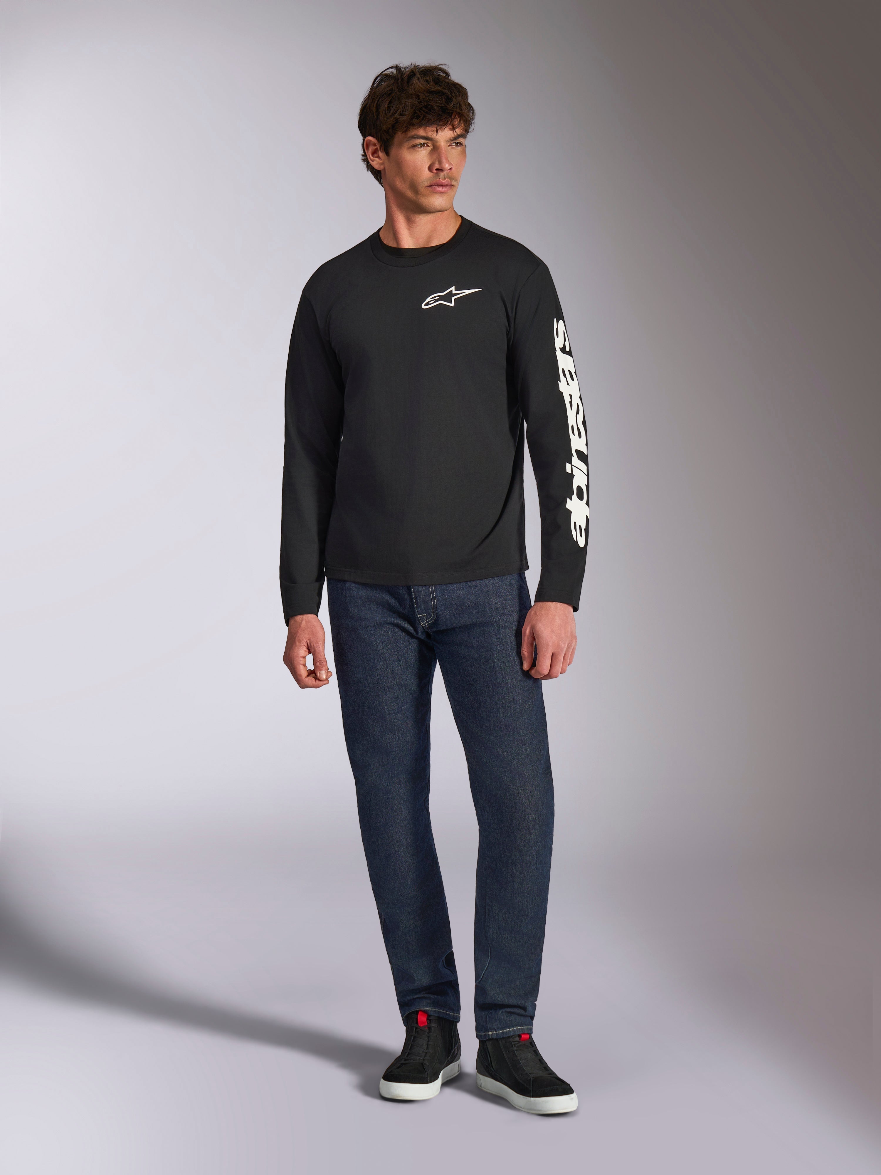 Alpinestars Trackside CSF Tee, T-shirt met lange mouwen, zwart en wit, gedragen met donkerblauwe jeans en zwarte sneakers, voorzien van wit Ride 4.0 logo op de borst en Alpinestars woordmerk op de mouw, casual motorsport-geïnspireerde streetwear