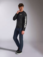 Alpinestars Trackside CSF Tee, T-shirt met lange mouwen, zwart, witte verticale Alpinestars branding op de mouw en logo op de borst, gedragen door mannelijk model met donkerblauwe jeans en sneakers, casual motorsport streetwear