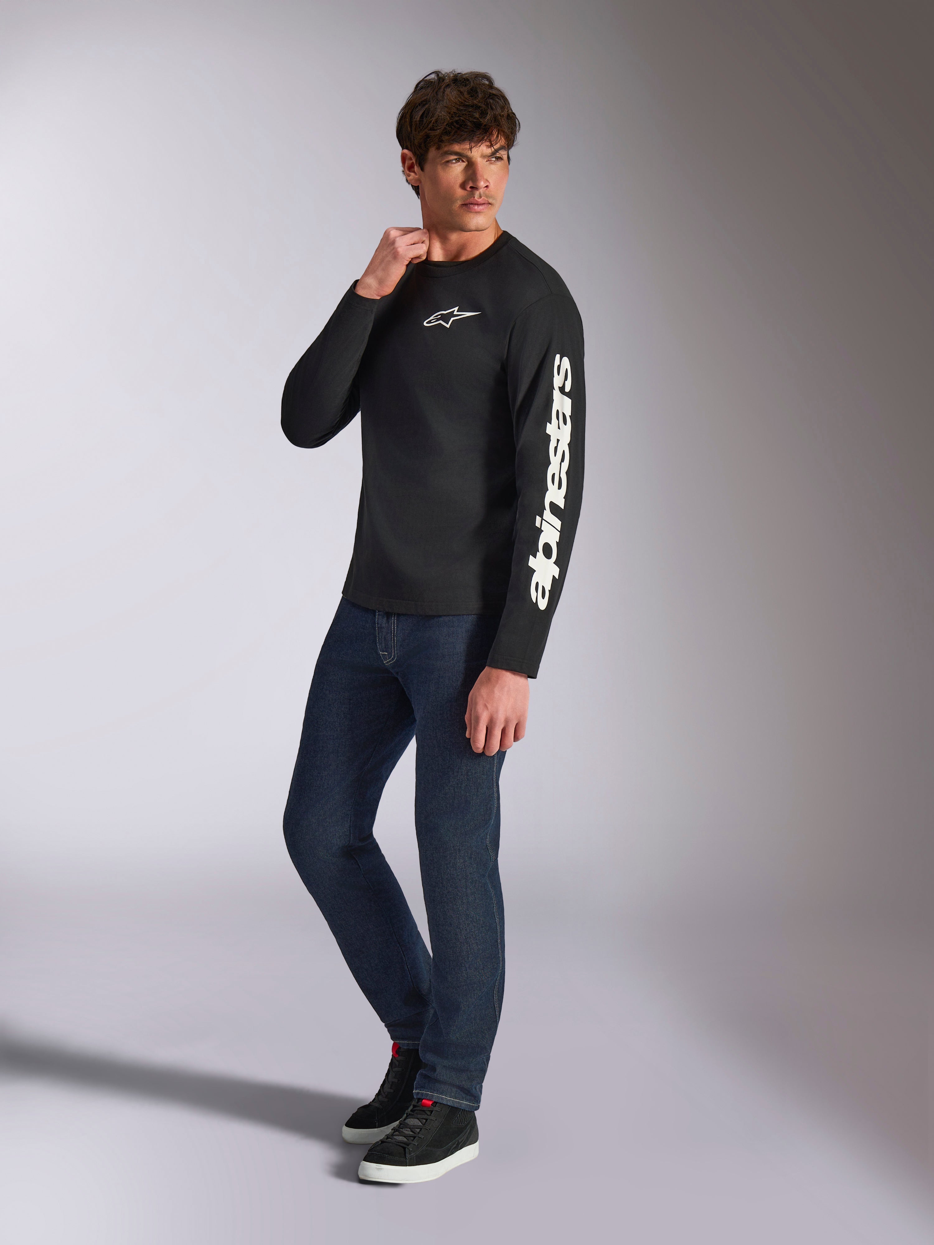 Alpinestars Trackside CSF Tee, T-shirt met lange mouwen, zwart, witte verticale Alpinestars branding op de mouw en logo op de borst, gedragen door mannelijk model met donkerblauwe jeans en sneakers, casual motorsport streetwear