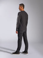 Alpinestars Trackside CSF T-shirt met lange mouwen, zwart, met groot grijs Alpinestars-logo op de linkermouw, gedragen door model met zwarte broek en casual sneakers, op motorsport geïnspireerde casual streetwear.