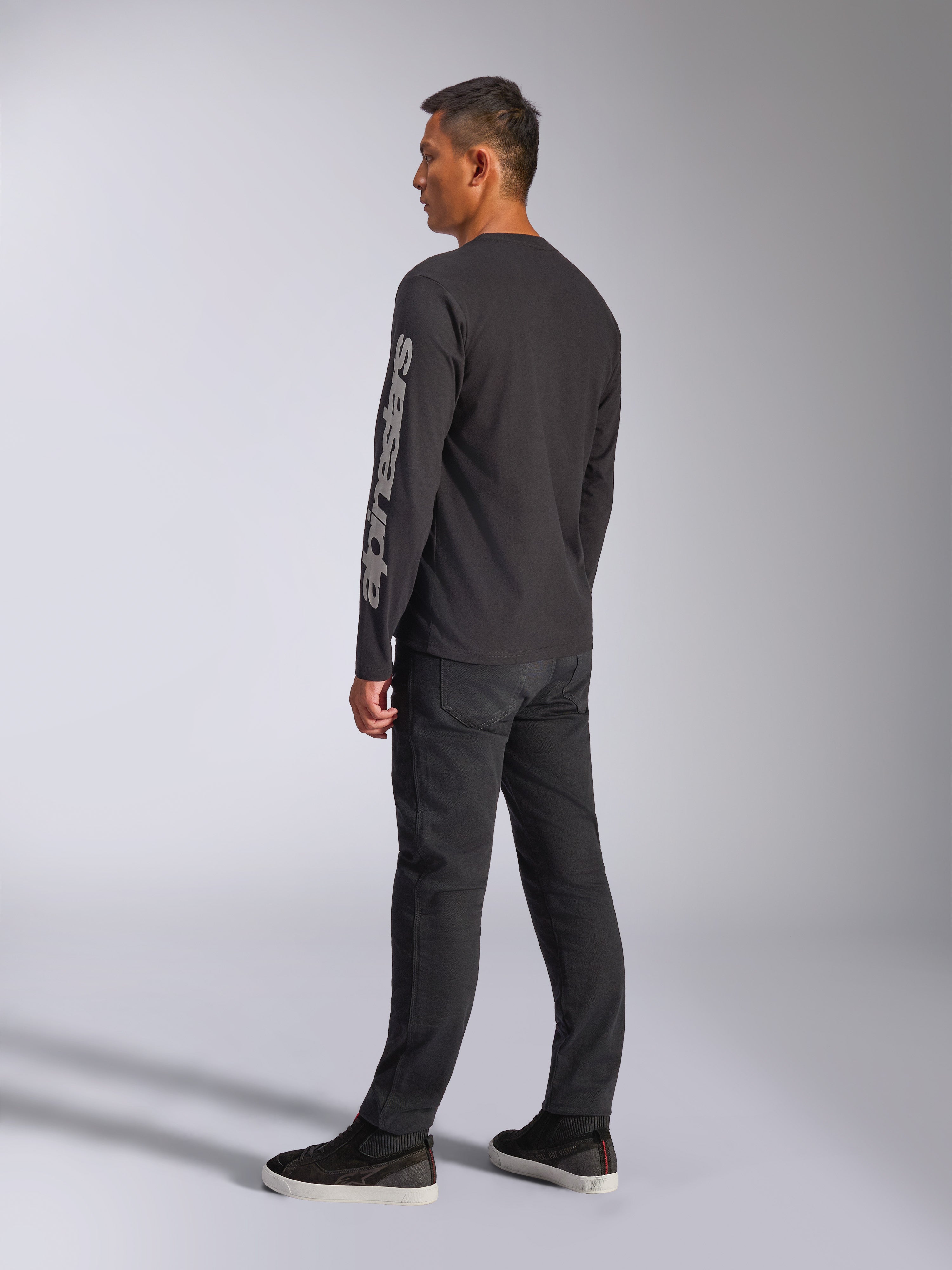Alpinestars Trackside CSF T-shirt met lange mouwen, zwart, met groot grijs Alpinestars-logo op de linkermouw, gedragen door model met zwarte broek en casual sneakers, op motorsport geïnspireerde casual streetwear.