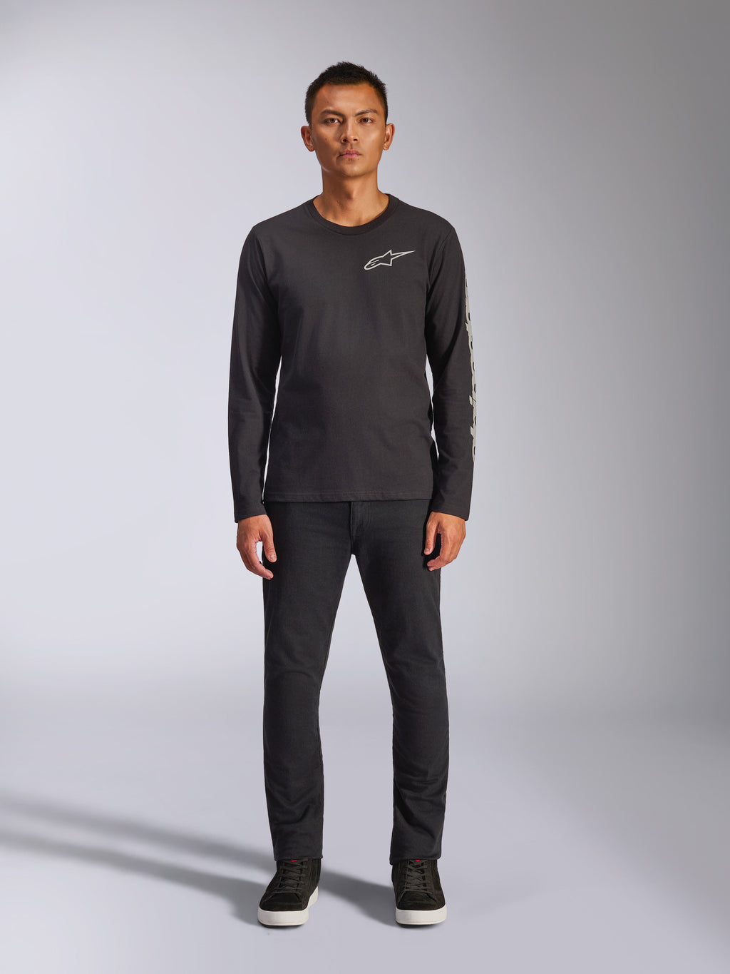 Alpinestars Trackside CSF Tee, T-shirt met lange mouwen, zwart, wit Alpinestars-logo op de borst en grafische branding op de mouw, gedragen met een zwarte broek en sneakers, casual motorsport-geïnspireerde streetwear