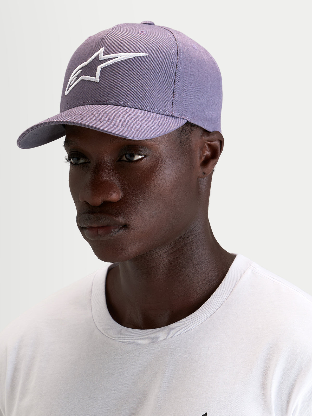 Alpinestars Casquette Snapback Ageless, casquette sportswear avec visière incurvée en violet poussiéreux et blanc, portée par un modèle présentant la couronne structurée et le logo Astars brodé en 3D