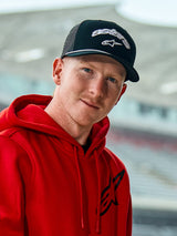Man wearing a red Alpinestars hoodie and the Agree Trucker Hat in Black, featuring white embroidered Astars logos, a white braided rope detail on the visor, and a breathable mesh back, casual lifestyle setting