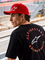 Alpinestars Corp Shift Multi Mütze, Rote Cap mit gebogenem Schirm und weißem 3D-Astars-Logo, getragen mit einem schwarzen Alpinestars T-Shirt mit „Racing Ahead“-Grafik, Lifestyle-Motorsport-Kulisse auf einer Rennstrecke.