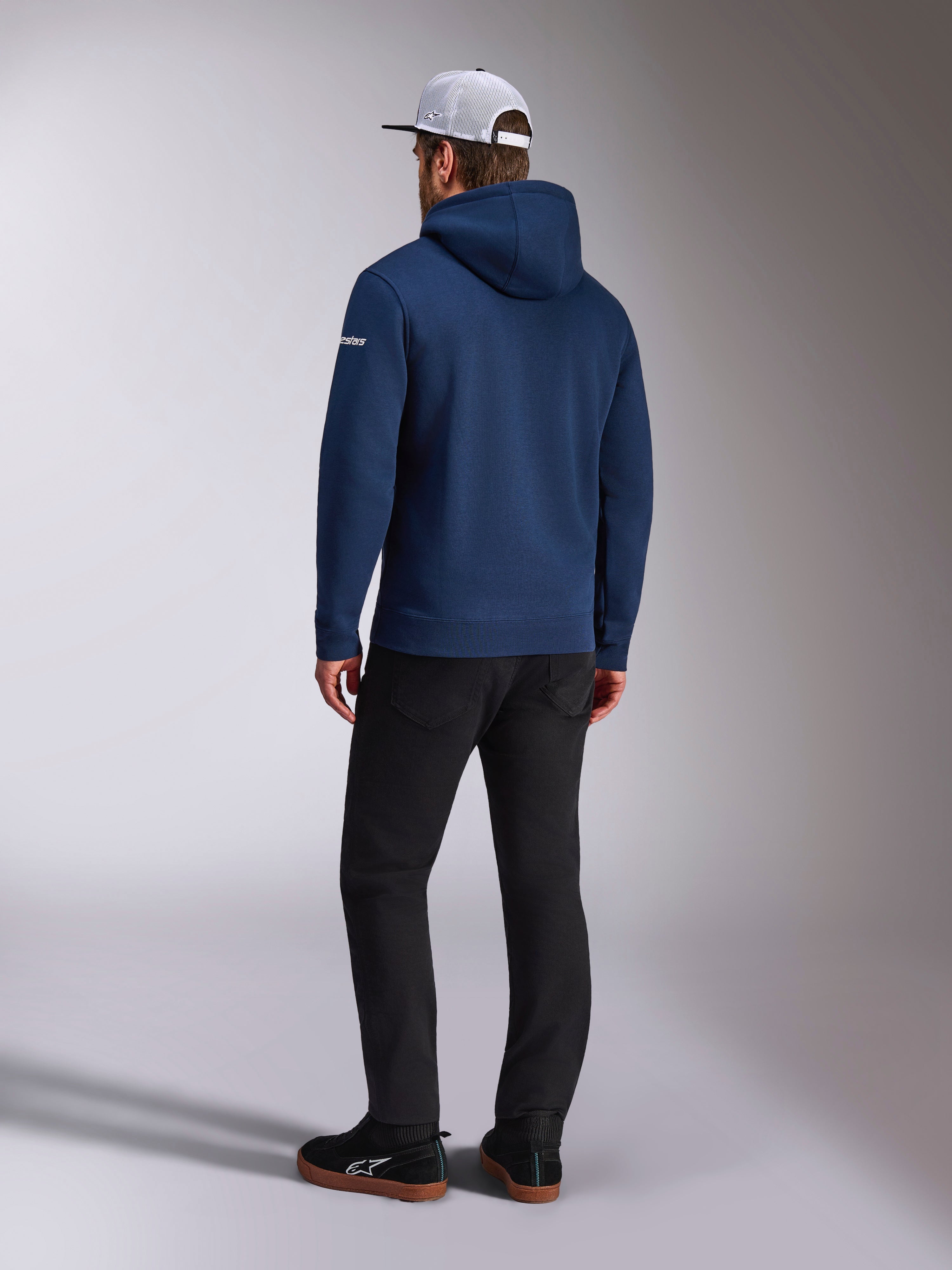 Alpinestars Frontal Hoodie, achteraanzicht van een marineblauwe pullover fleece hoodie, wit woordlogo op de mouw, gecombineerd met een zwarte broek en een witte mesh truckerpet, casual op motorsport geïnspireerde streetwear