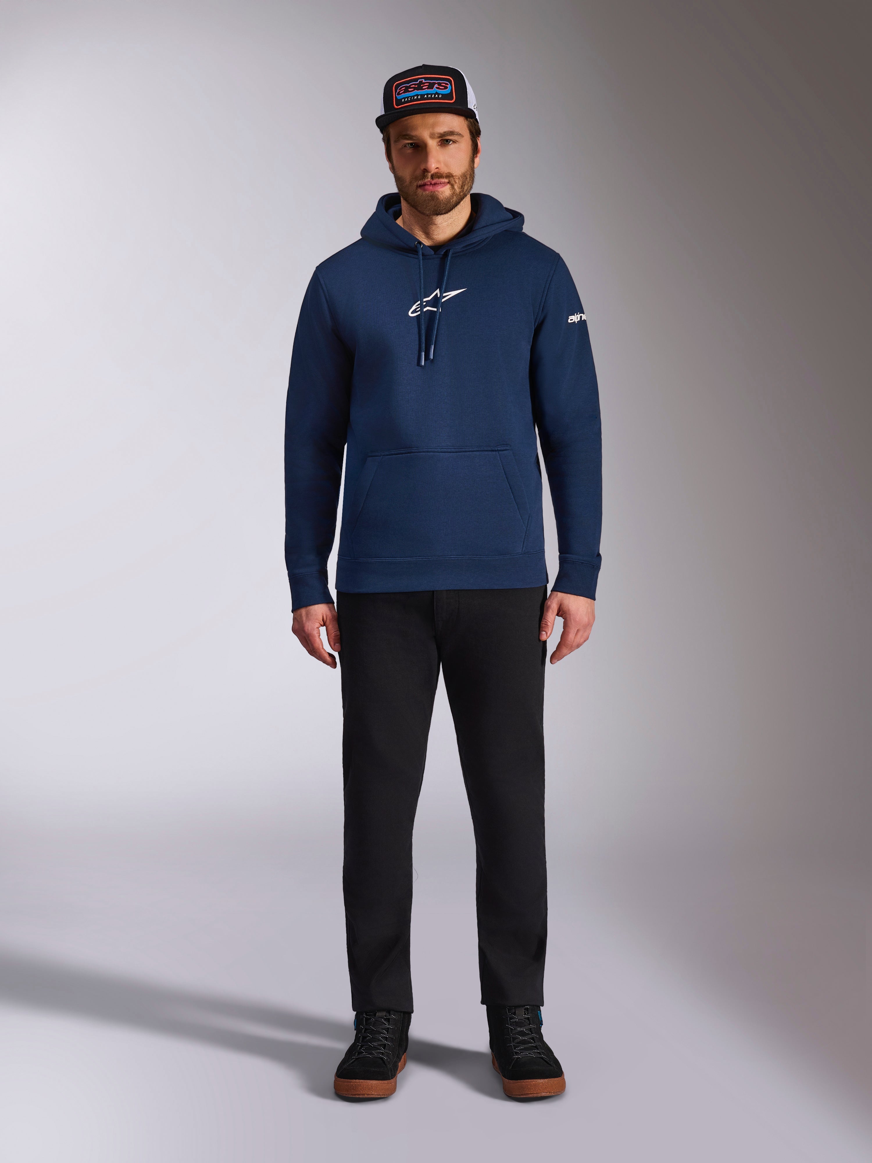 Mannelijk model draagt de Alpinestars Frontal Hoodie in Navy, fleece pullover met een wit Astars-logo op de borst en woordlogo op de mouw, voorzien van een kangoeroezak en capuchon met trekkoord, gecombineerd met een zwarte broek en een pet met logo, casual motorsport streetwear