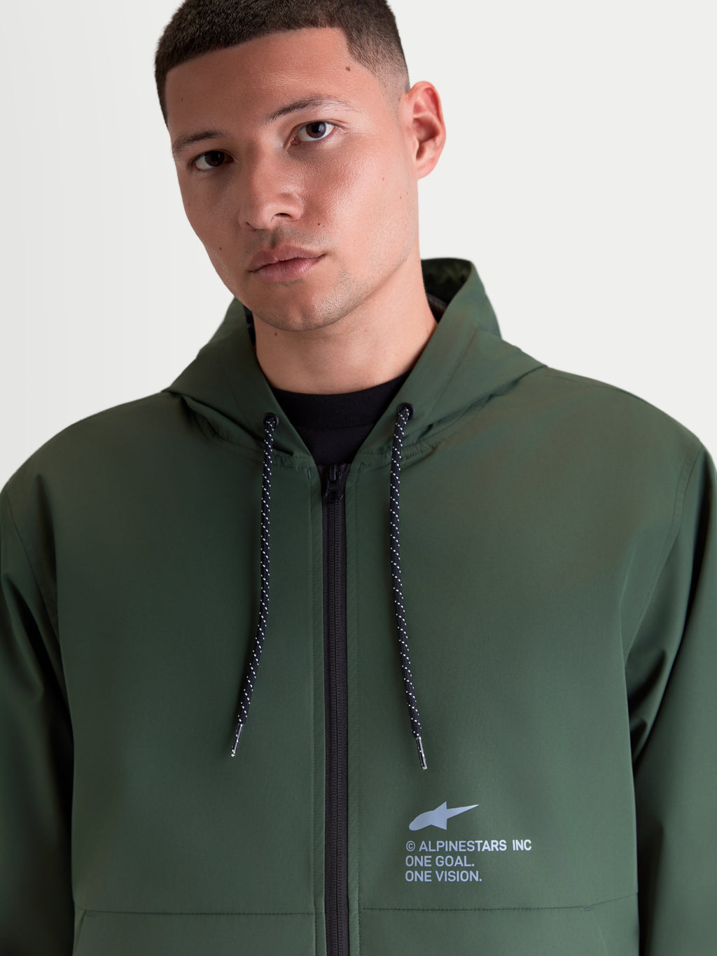 Alpinestars Speeder Resolve, veste coupe-vent de sportswear légère pour homme, vert, dotée d'une capuche réglable avec cordons mouchetés et marquage Alpinestars sur la poitrine, porté par un modèle.