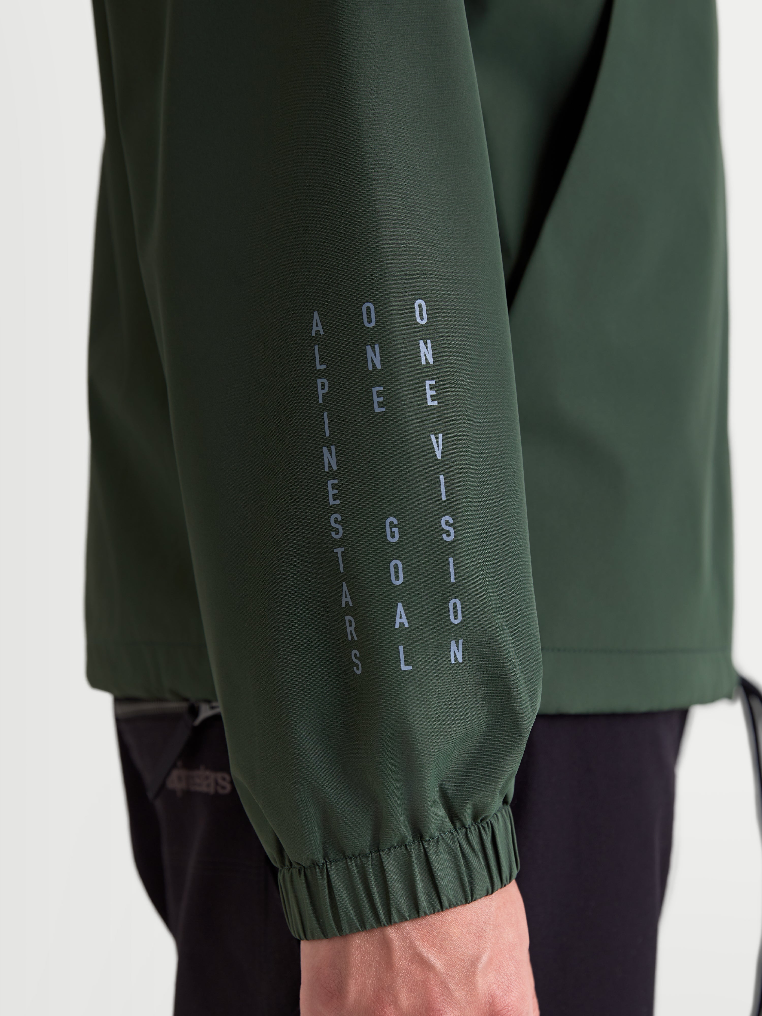 Alpinestars Speeder Resolve, veste coupe-vent légère de sportswear, vert pin, vue rapprochée de la manche avec marquage vertical Alpinestars, devise One Goal One Vision et poignet élastique.