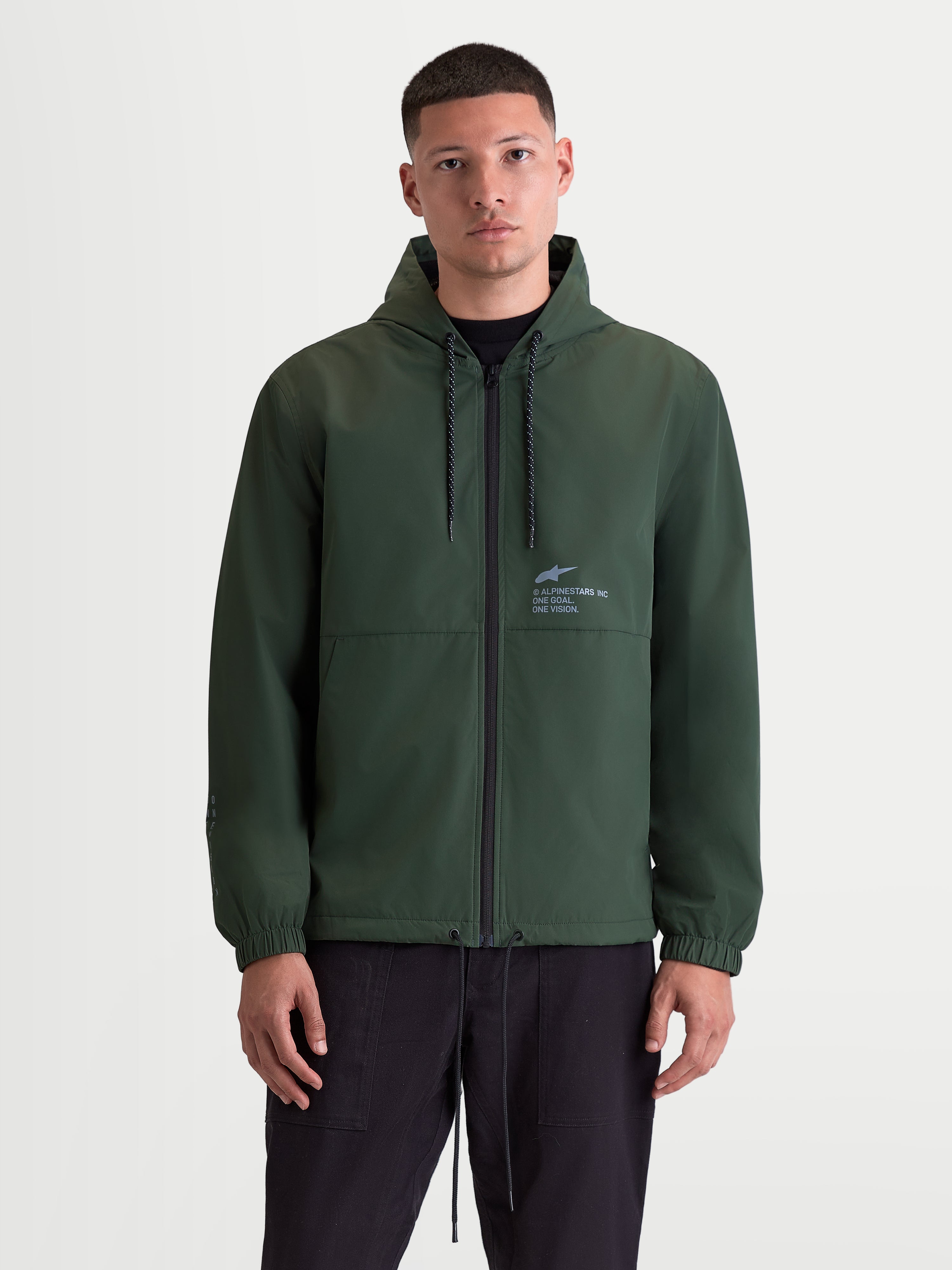 Alpinestars Veste Coupe-Vent Speeder Resolve, veste de sportswear pour usage urbain, veste à capuche pour homme vert sapin, intérieur doublé en mesh, capuche réglable avec cordon de serrage et fermeture éclair frontale, marquage Alpinestars sur la poitrine, vue de face sur modèle.