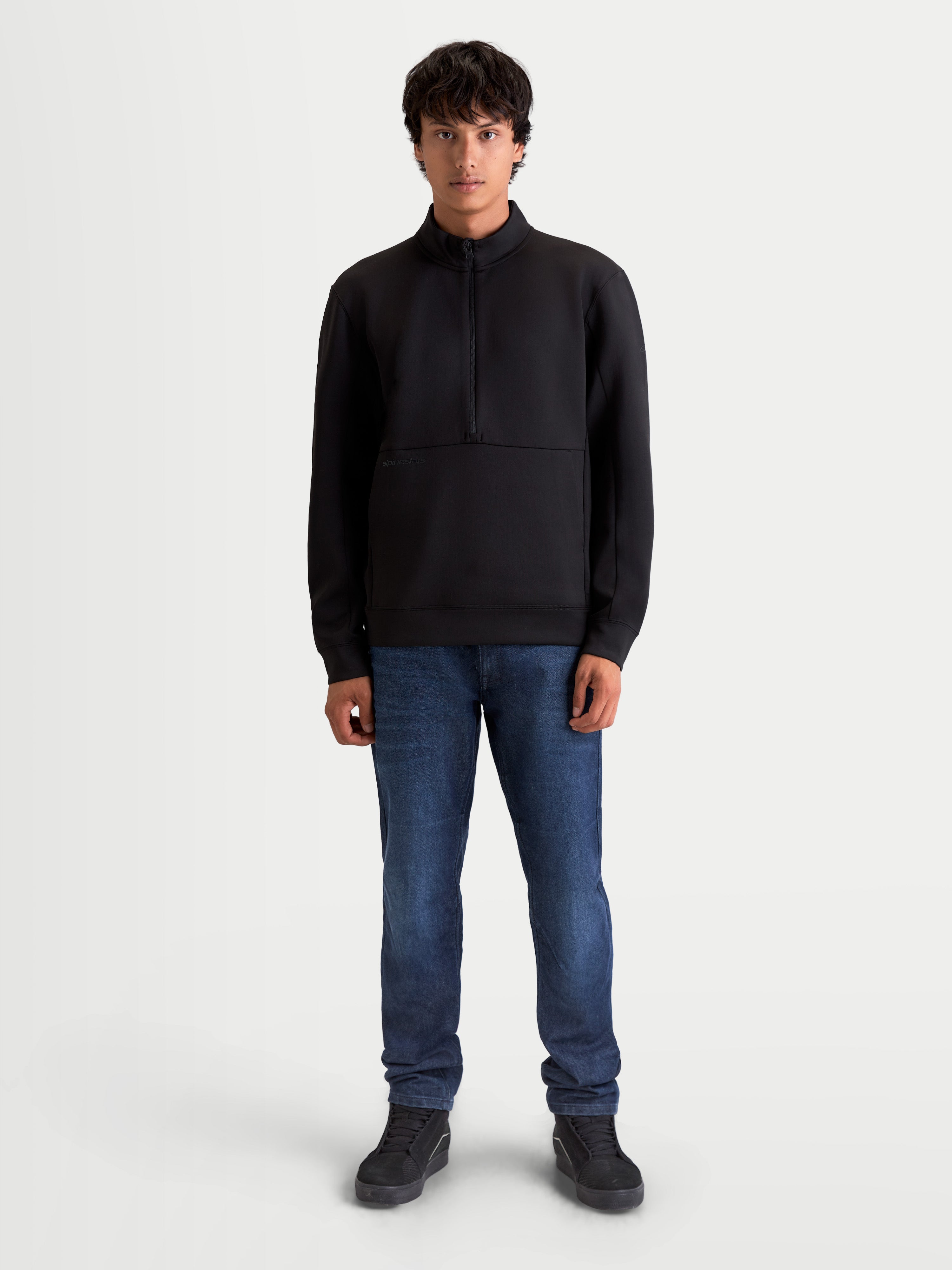 Alpinestars Sweat Half-Zip Apera, pull sportswear pour homme, noir, avec fermeture demi-zippée et poche kangourou, style inspiré du sport automobile pour un confort décontracté, porté par un modèle avec un jean bleu.