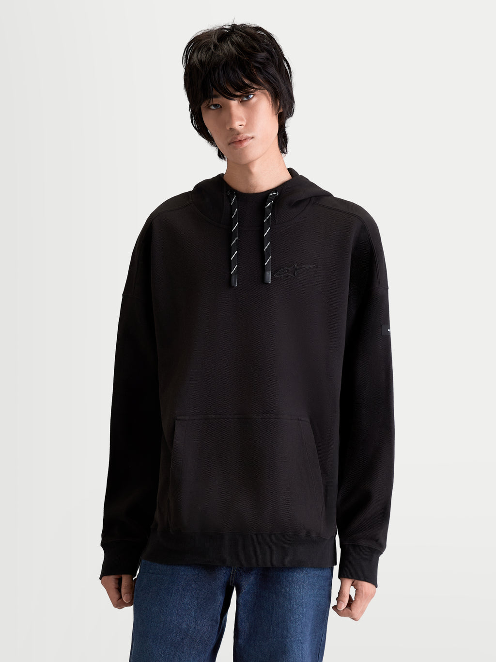 Alpinestars sweat à capuche Textured Oversize Camino, vêtement de style sportswear unisexe, noir, polaire en maille sergée texturée avec col haut, poche kangourou et logo brodé ton sur ton sur la poitrine, porté par un modèle.