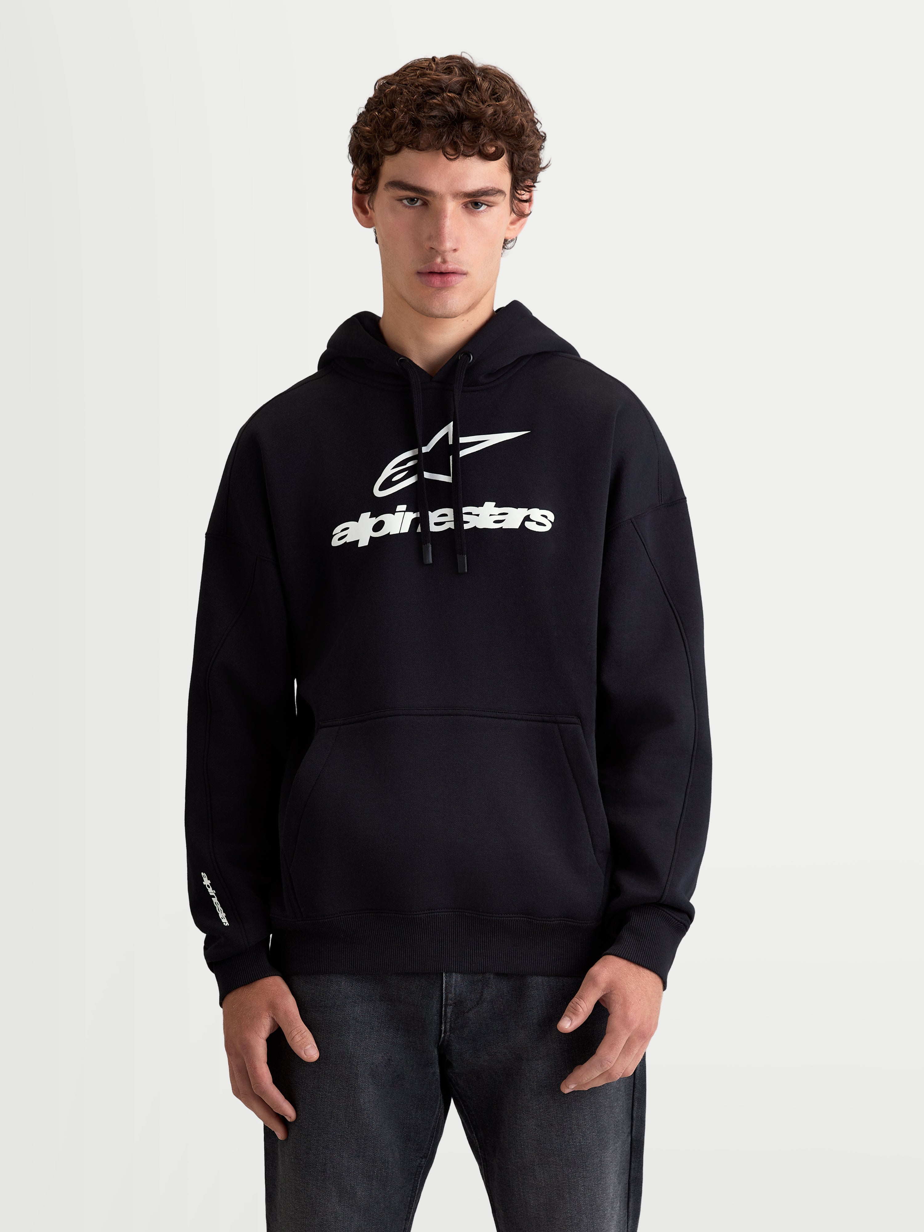 Alpinestars sweat à capuche Prelude Oversized, sweat à capuche de sportswear en polaire pour homme, noir et blanc, porté par un modèle, coupe oversized avec poche kangourou et logos en relief sur la poitrine et la manche, style décontracté