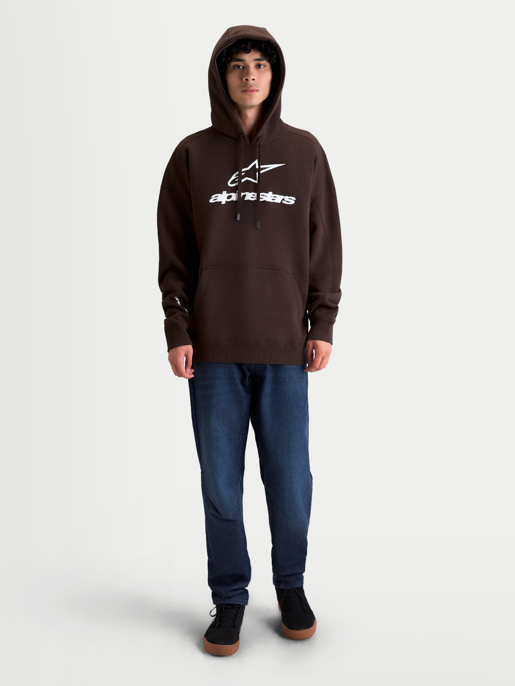 Alpinestars Prelude Oversized Hoodie, bruine sportswear fleece pullover, unisex, katoenrijk materiaal, met een ontspannen oversized pasvorm, wit puffprint-logo op de borst, kangoeroezak en gevoerde capuchon, getoond op model.