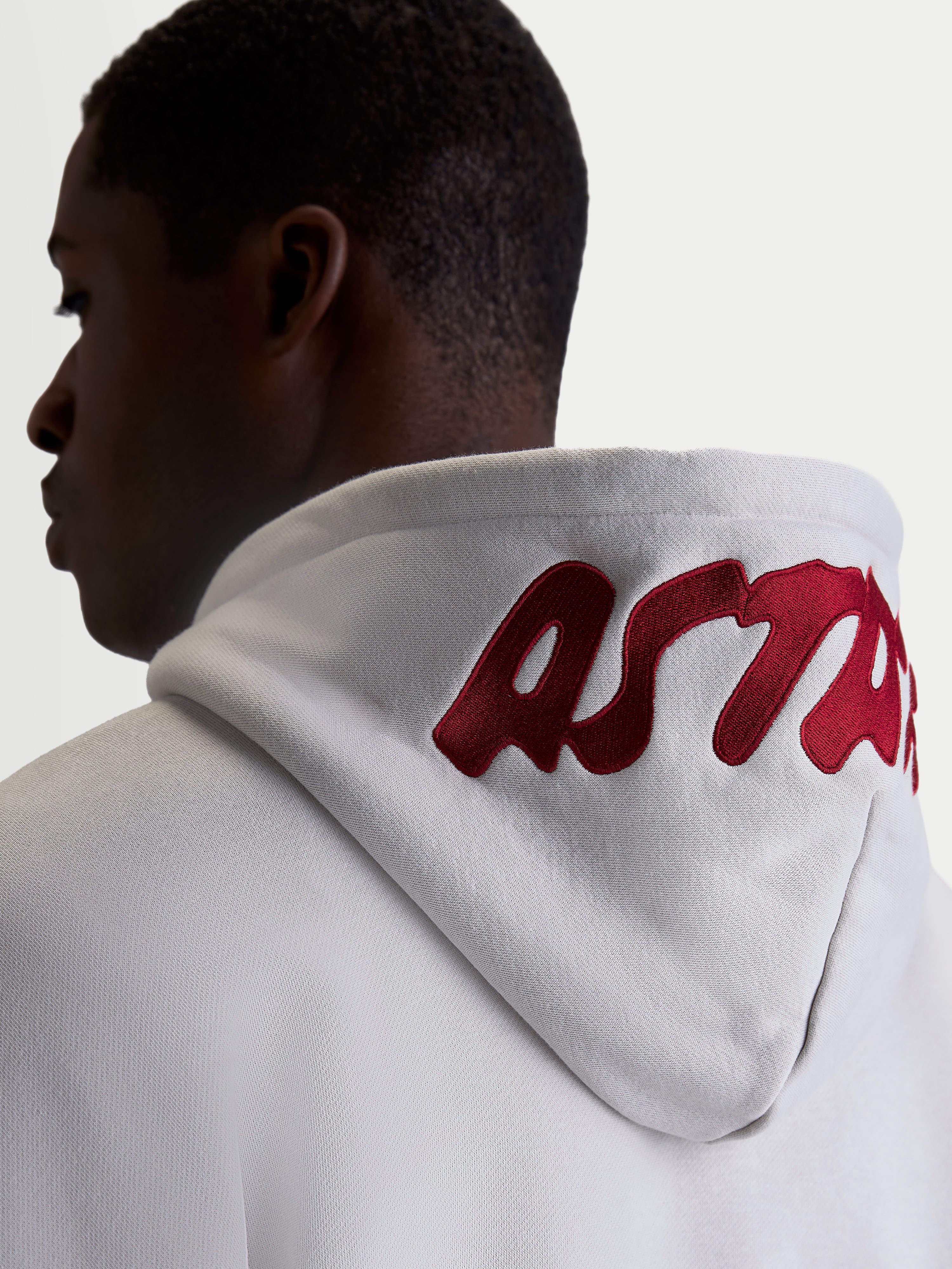 Alpinestars Sweat à capuche Pioneer Oversized, polaire de sportswear décontractée pour homme et femme, gris ciment avec logo brodé rouge sur la capuche, vue de dos sur modèle mettant en avant la coupe oversized et le marquage premium