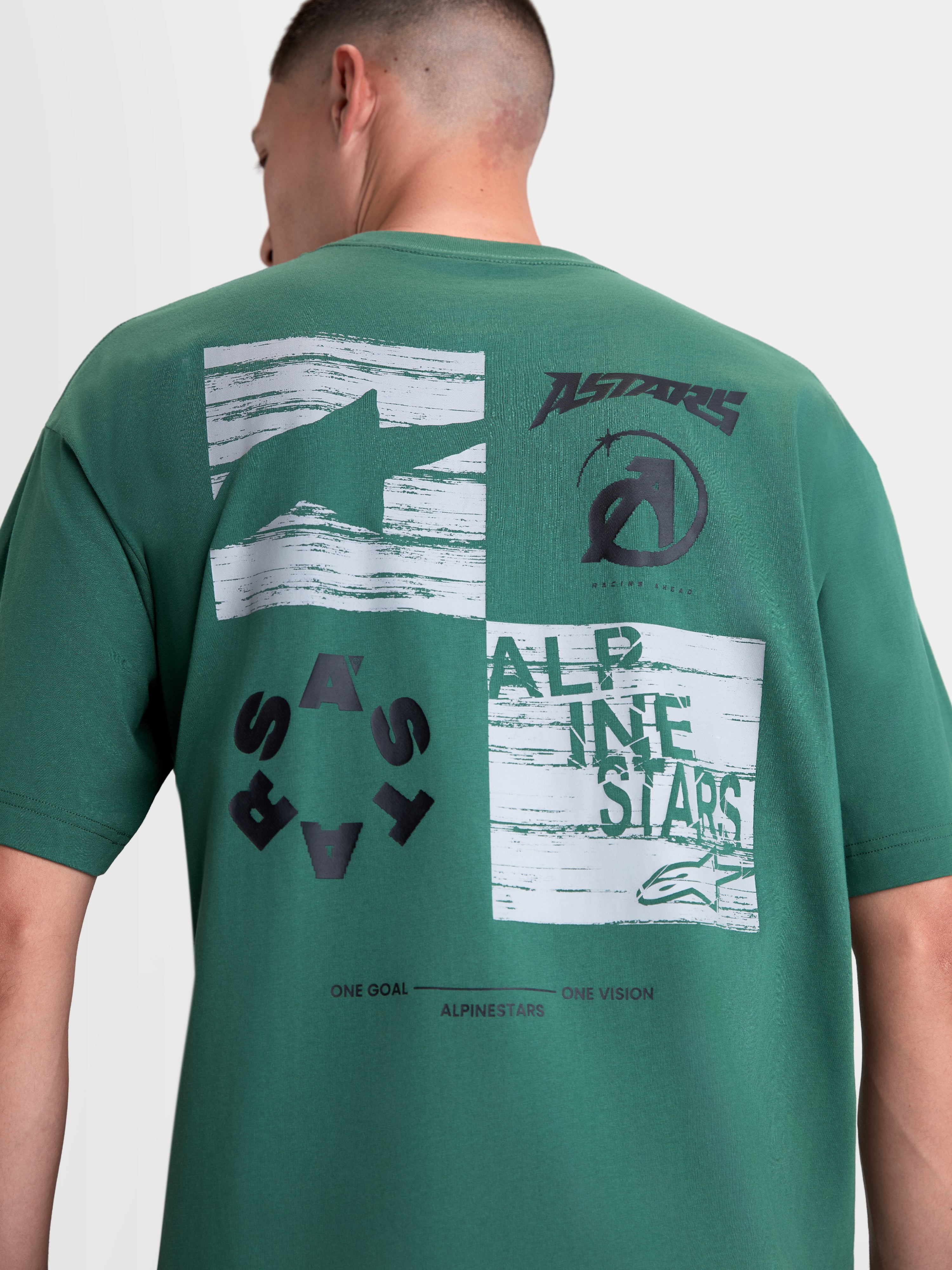 Alpinestars Checkback Oversized CSF Tee, Green Sportswear Short Sleeve T-Shirt, Men's and Women's Casual Wear, Back View on Model. Features a prominent black and white back checkerboard graphic with Astars logos and 'One Goal One Vision' text. Constructed from a soft cotton-poly blend with an oversized fit for comfort and motorsport-inspired style.