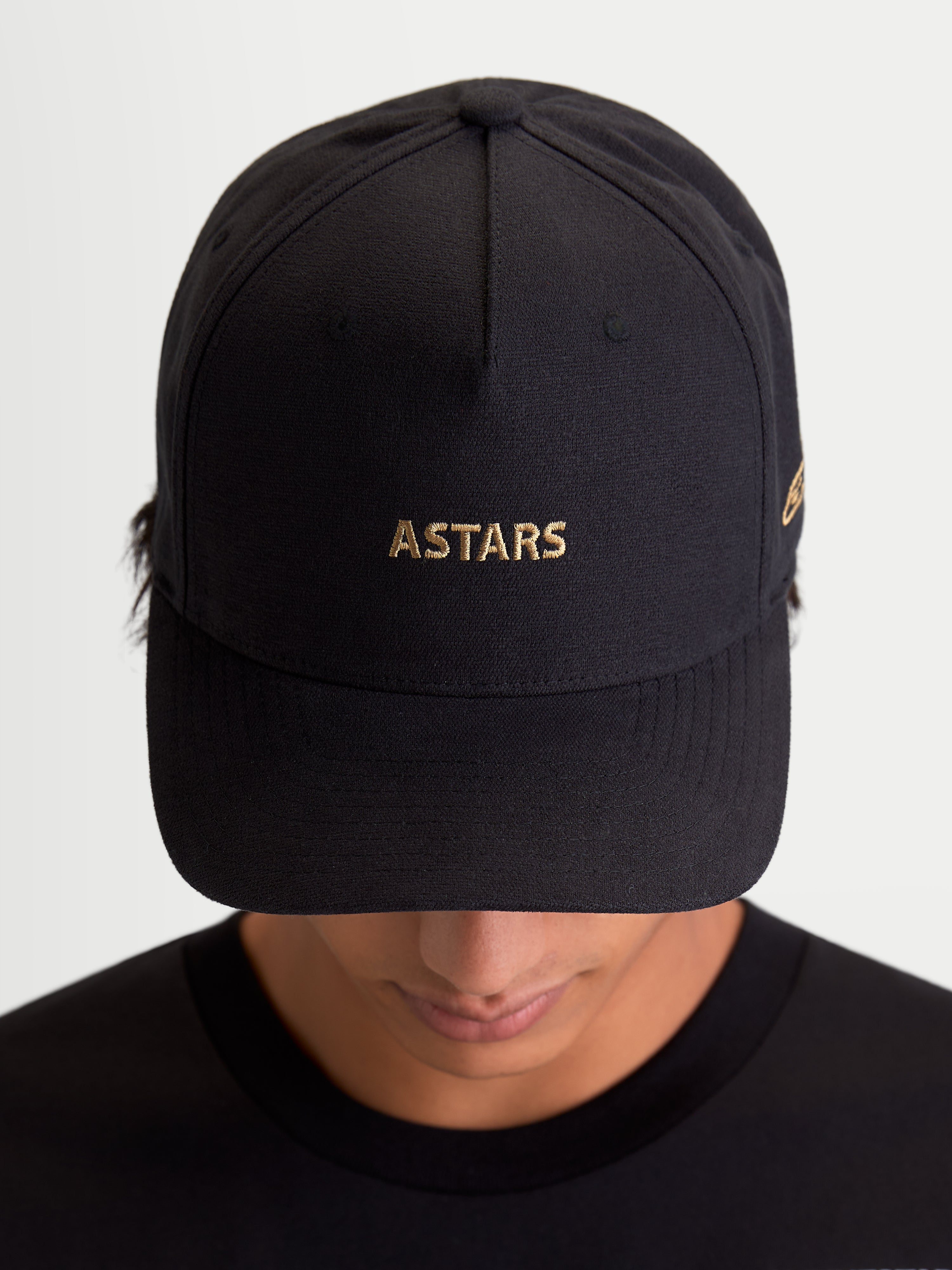 Alpinestars Casquette County Canvas, casquette de sportswear noire avec logo ASTARS doré brodé, visière incurvée et couronne structurée, tissu oxford, vue de face sur un modèle, style inspiré du sport automobile.