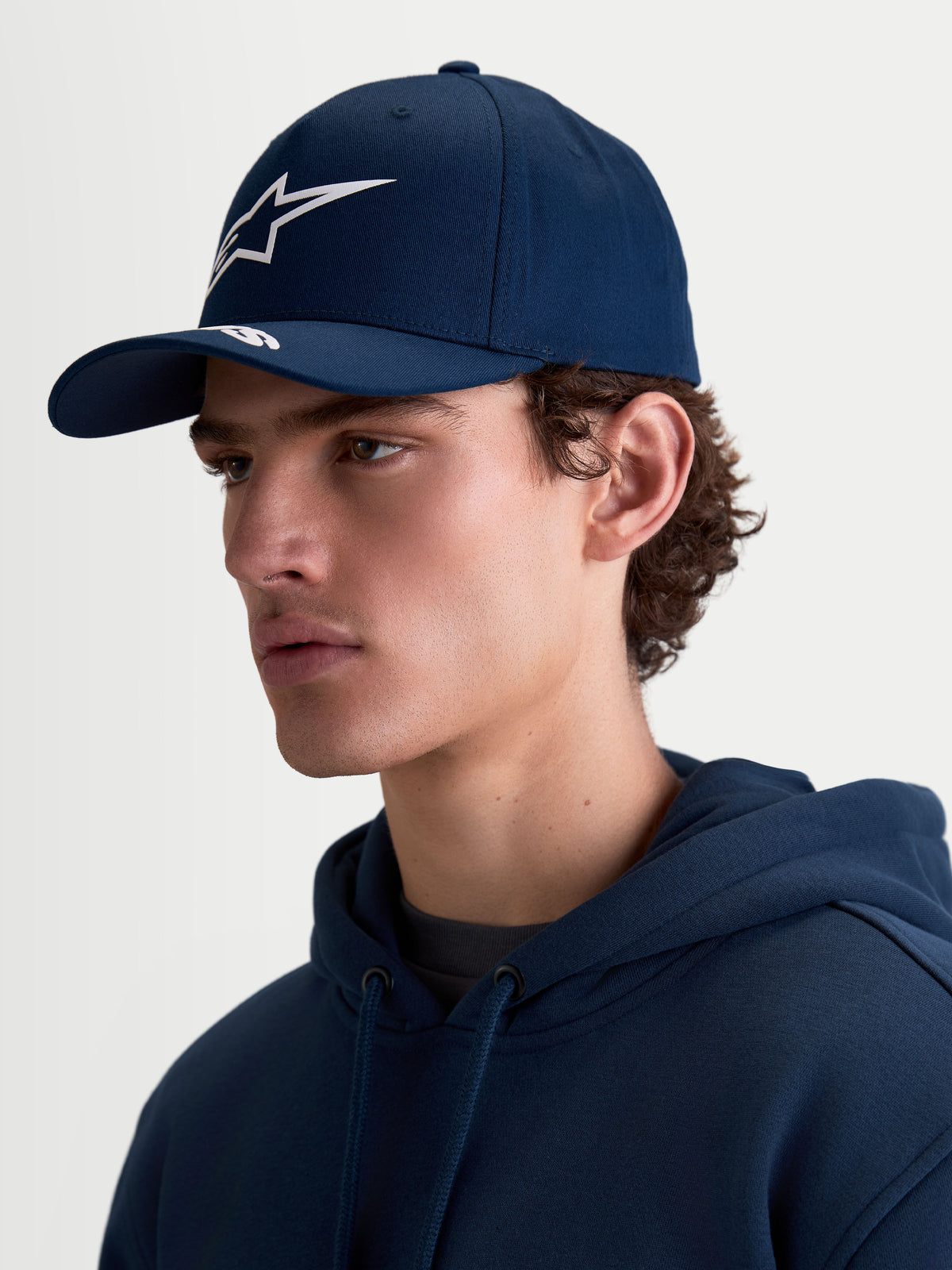 Alpinestars Casquette Arclight, casquette de sportswear bleu marine, visière incurvée et couronne structurée, logo Astars blanc imprimé en relief, vue de profil sur mannequin montrant la coupe motorsport en sergé de coton.