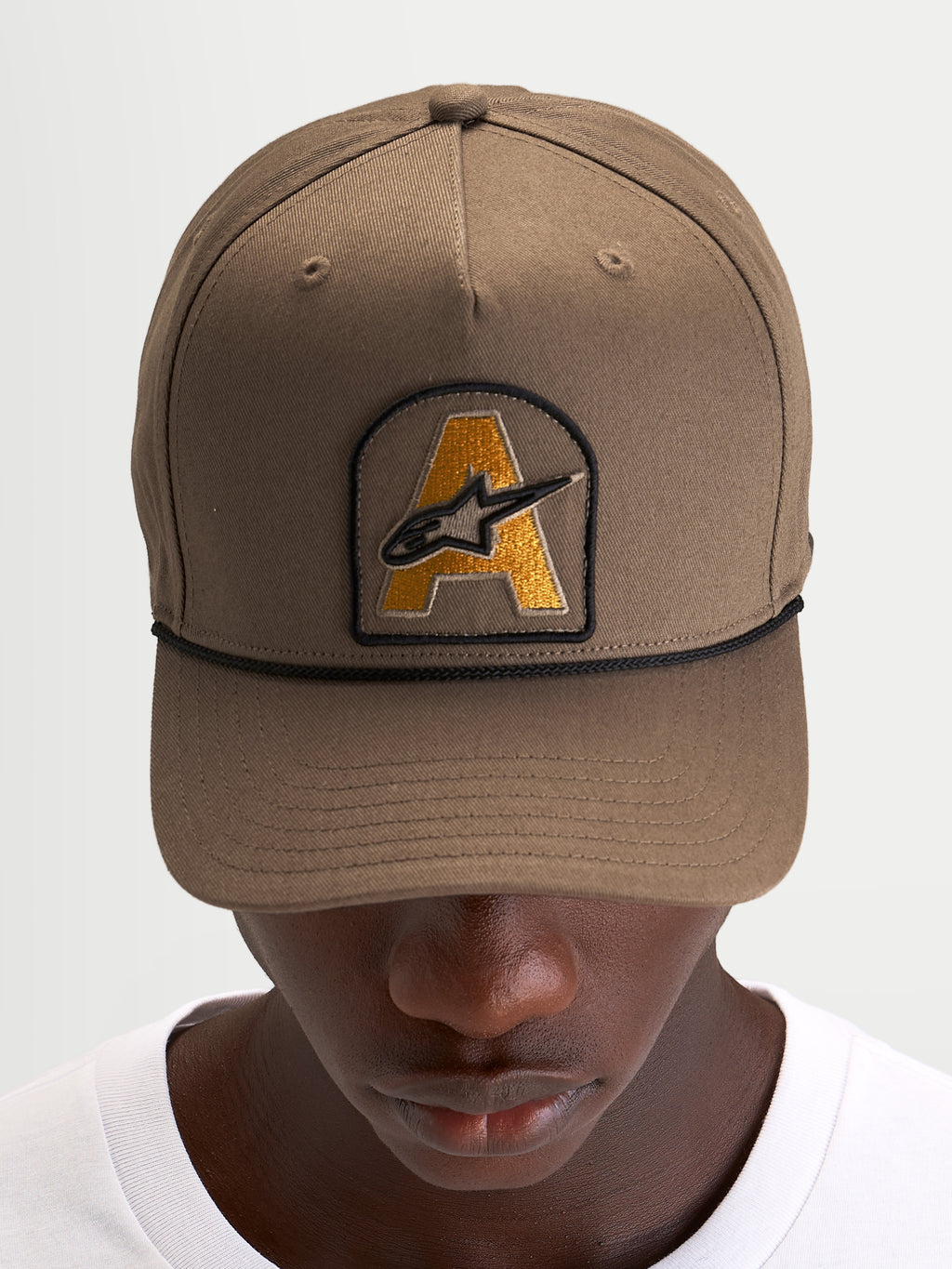 Alpinestars Casquette Acme Snapback, casquette de sport marron avec visière courbée, écusson brodé du logo Astars à l'avant et cordon noir, portée par un modèle