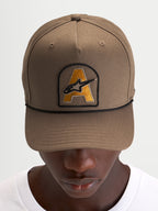 Alpinestars Casquette Acme Snapback, casquette de sport marron avec visière courbée, écusson brodé du logo Astars à l'avant et cordon noir, portée par un modèle
