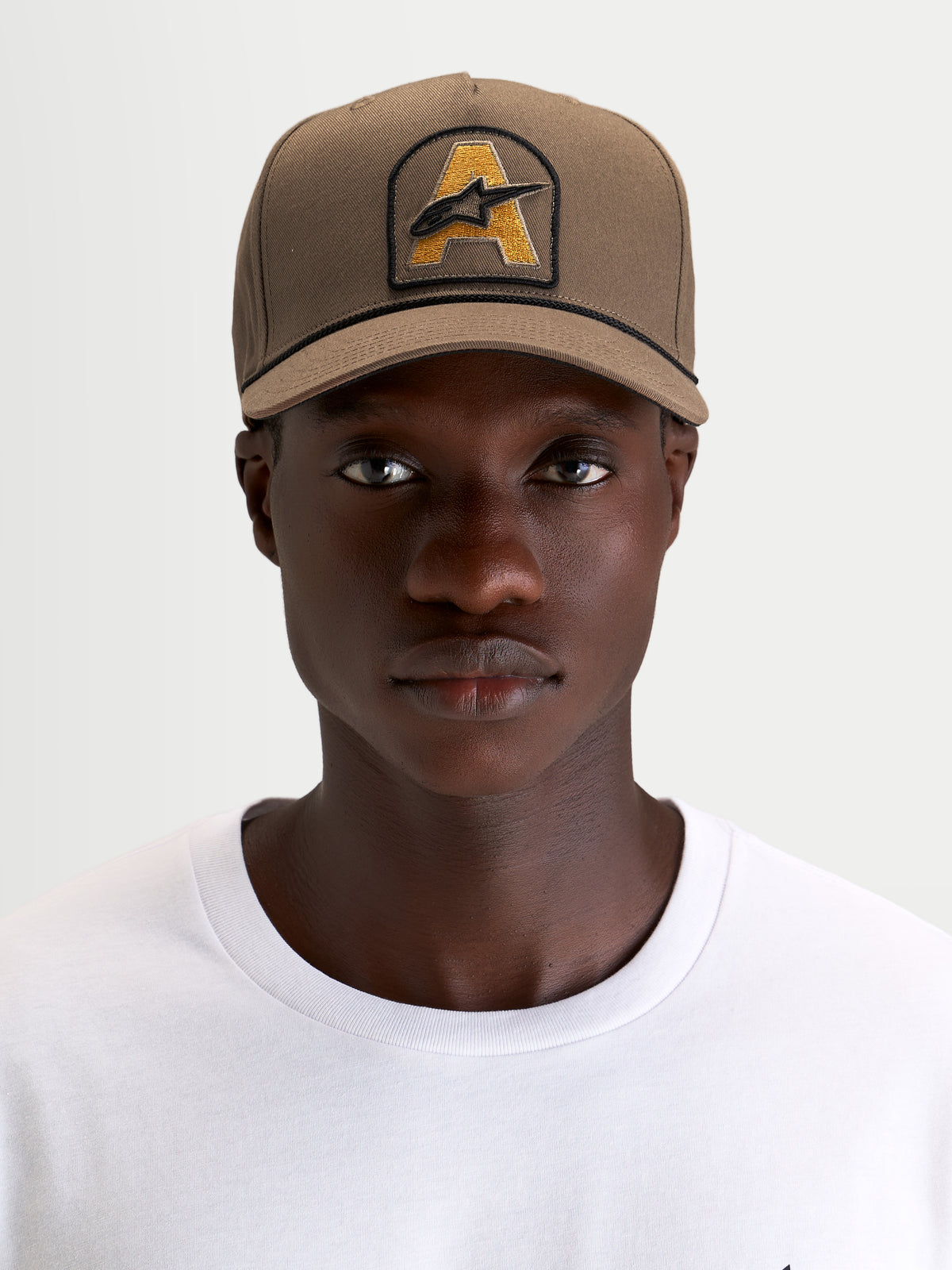 Alpinestars Acme Snapback pet, bruine sportswear pet gedragen door een model, met gestructureerde kroon, gebogen klep en Astars-logopatch op de voorkant met koordrand