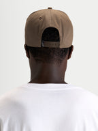 Alpinestars Casquette Acme Snapback, casquette sportswear marron portée à l'envers par un modèle, montrant la fermeture snapback réglable et l'étiquette arrière