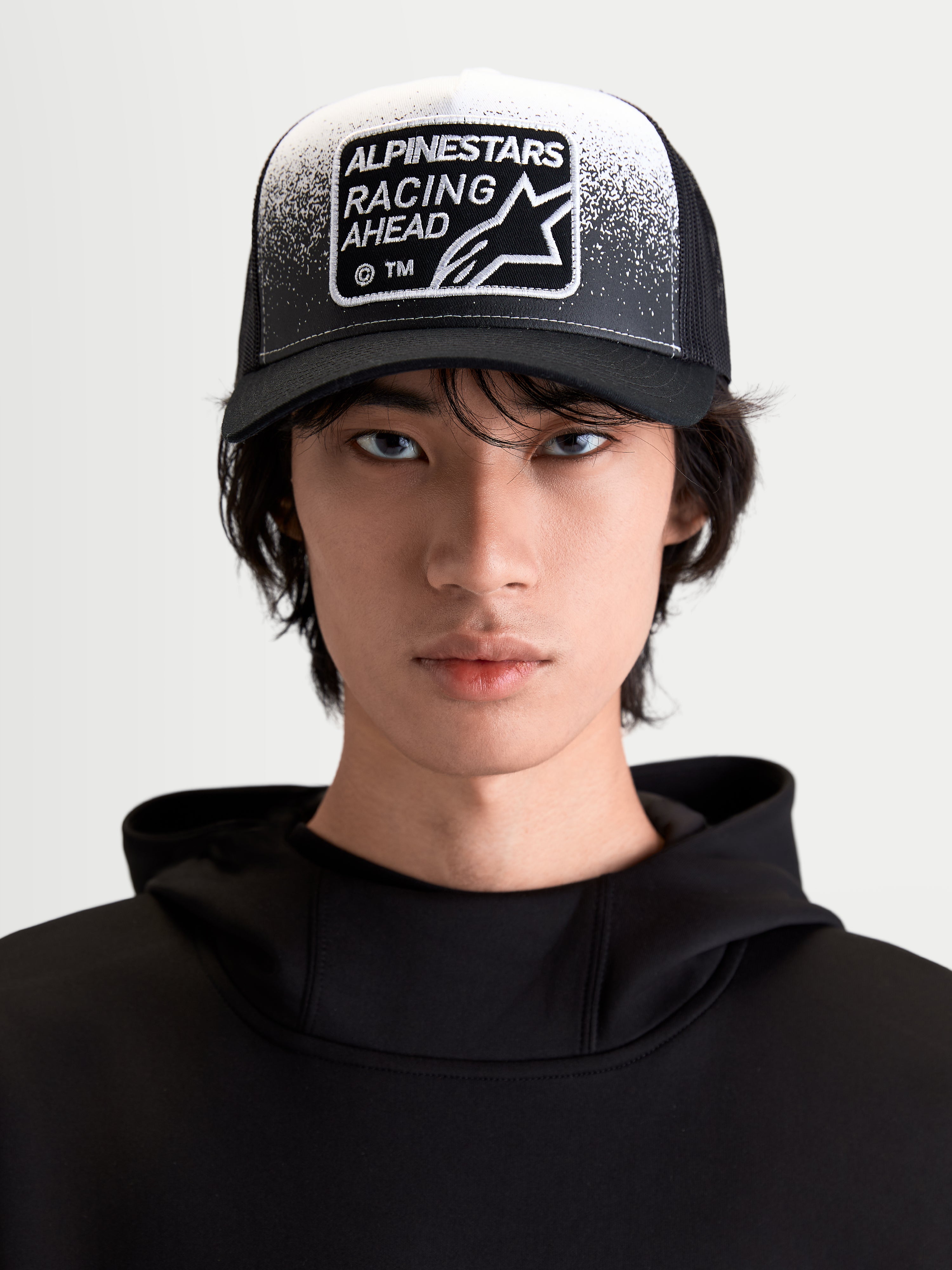 Alpinestars casquette Trucker Gravel, casquette de sportswear lifestyle, noire et blanche, vue de face sur un mannequin avec visière courbée, arrière en maille respirante et écusson tissé Racing Ahead avec détails effet gravier