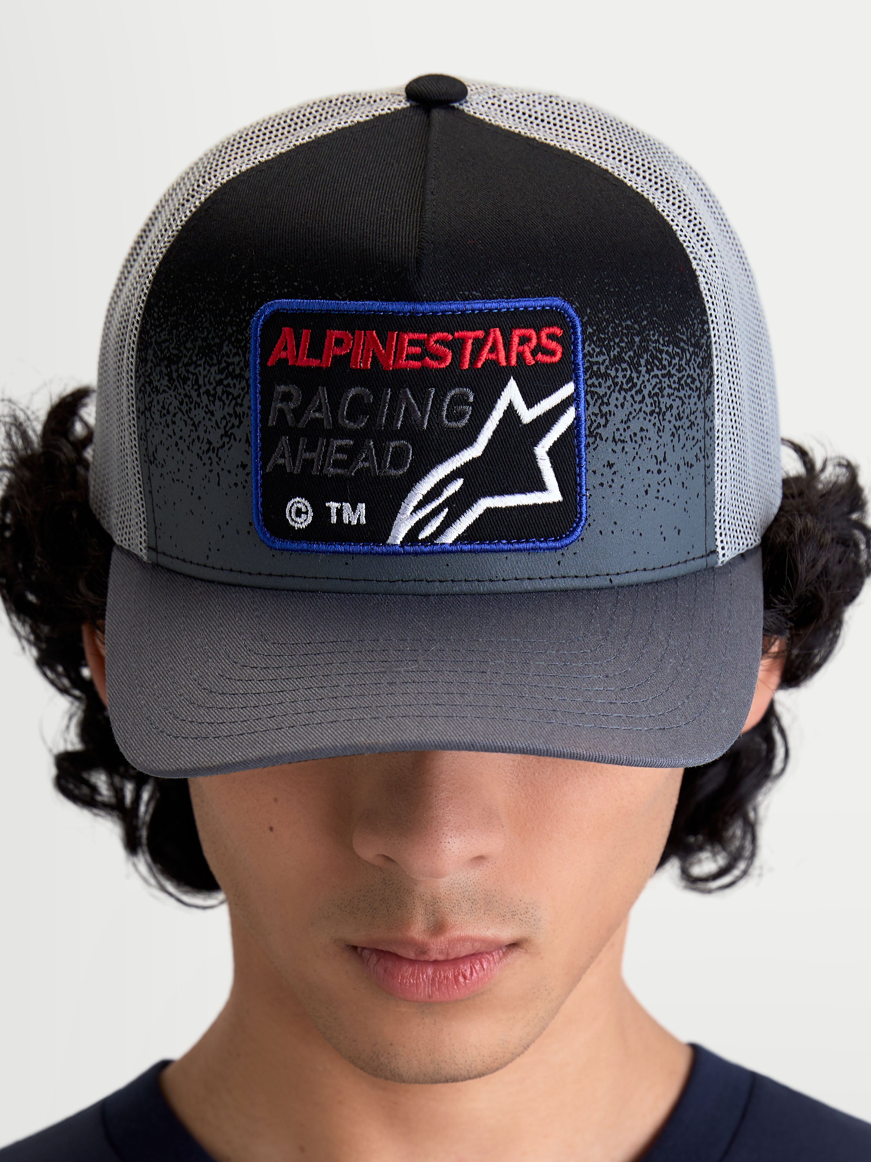 Alpinestars Gravel Trucker Hat, Sportswear Mesh Cap, Dark Gray and Black, featuring a curved bill and a woven front patch with 'Racing Ahead' text and Astars logo, on-model front view.