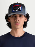 Alpinestars Gravel Trucker Hat, casual sportswear mesh back cap for men and women, Dark Grey and Black, shown on a model. Features a structured crown, curved bill, and a woven front patch with Alpinestars Racing Ahead branding and gravel-texture detailing.
