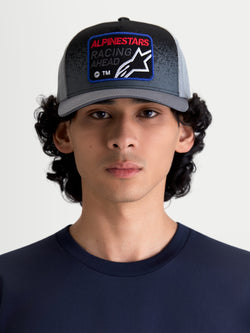 Alpinestars Gravel Trucker Hat, casual sportswear mesh back cap for men and women, Dark Grey and Black, shown on a model. Features a structured crown, curved bill, and a woven front patch with Alpinestars Racing Ahead branding and gravel-texture detailing.