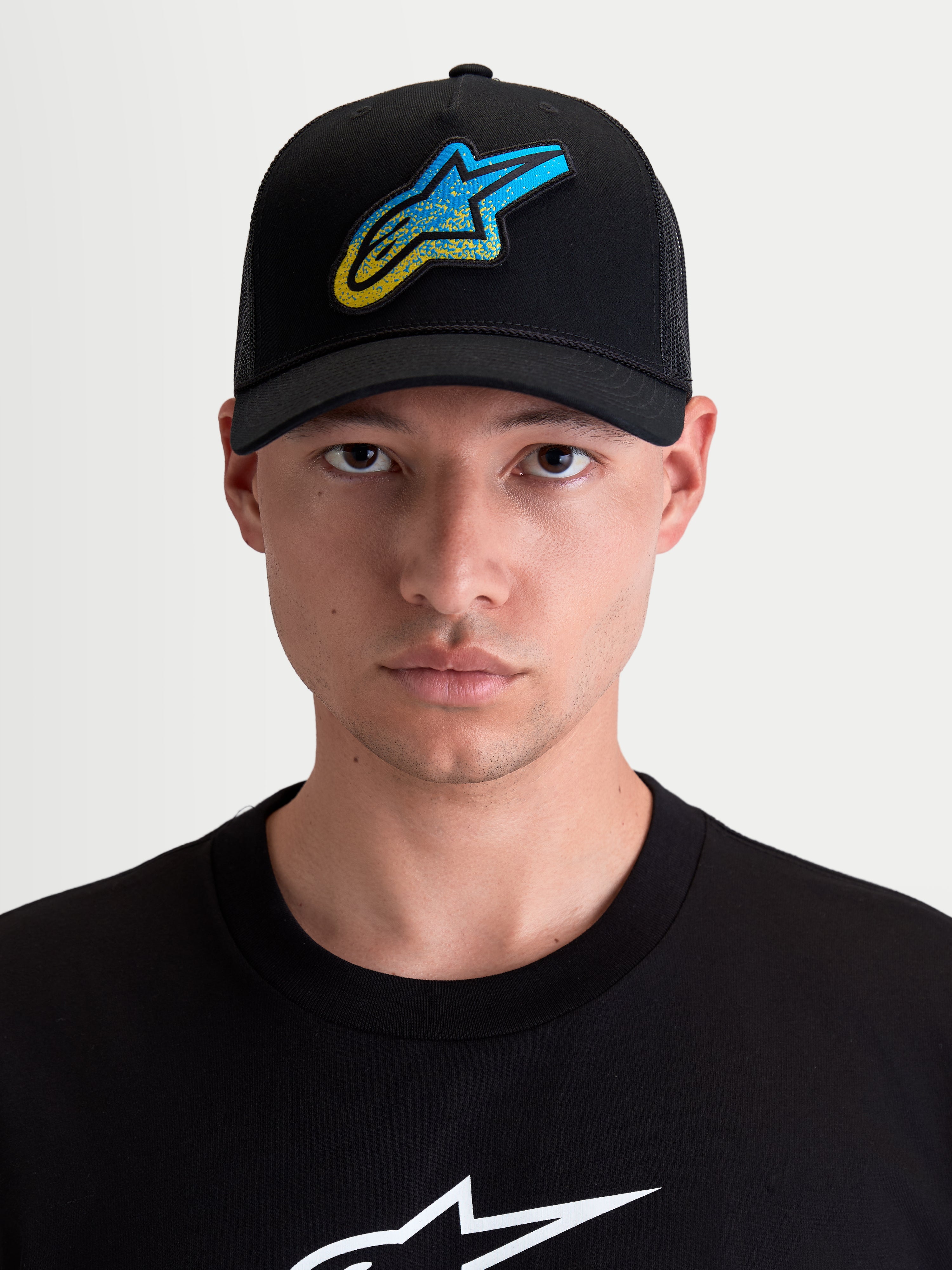 Alpinestars Meteorite trucker pet, sportswear cap met gebogen klep voor heren en dames, zwart met een blauw en geel Astars-logo met grindtextuur, twill voorzijde en poly mesh achterkant, vooraanzicht op model.