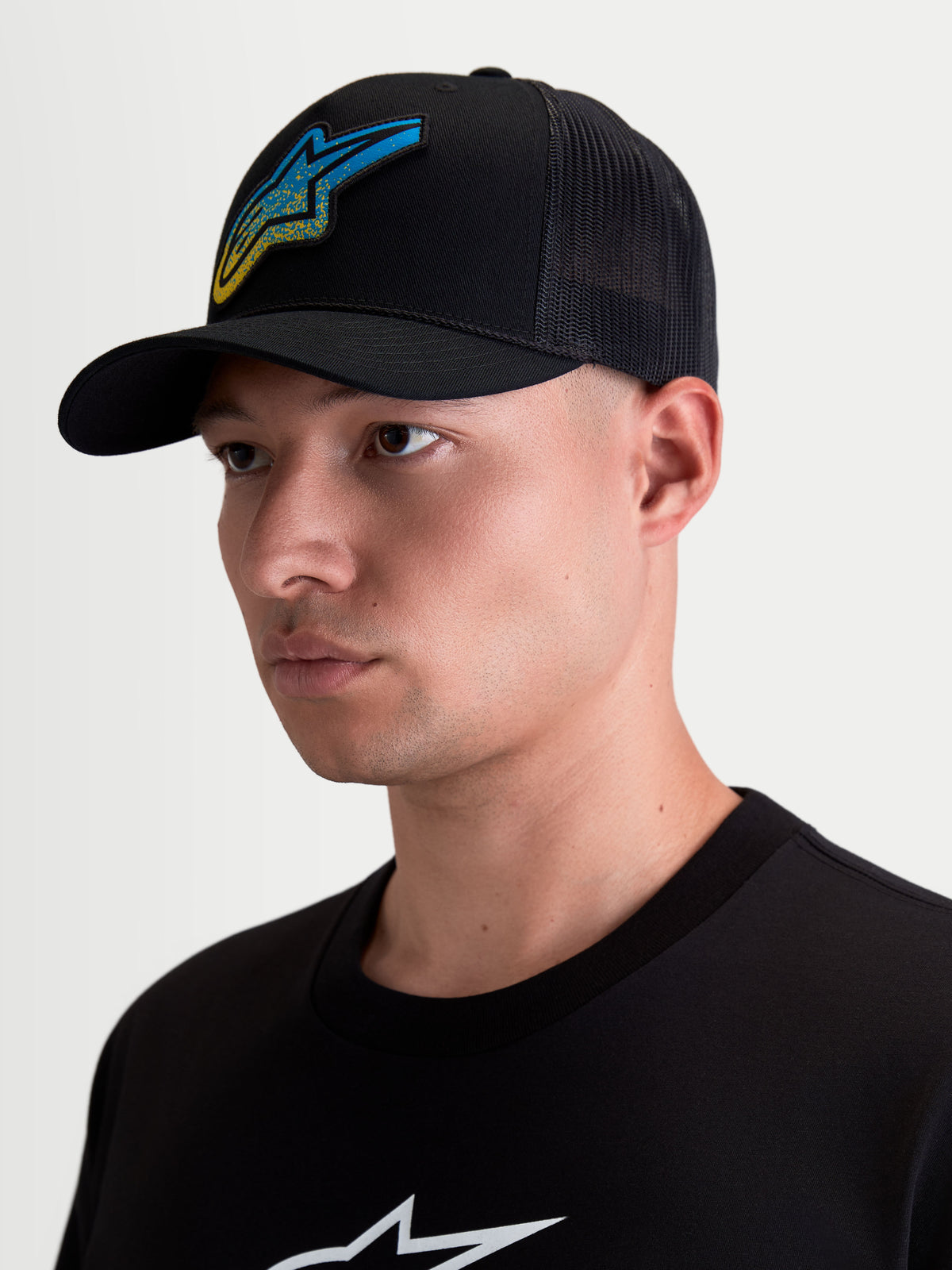 Alpinestars Meteorite Trucker pet, casual sportswear lifestylepet met mesh-achterzijde voor heren, zwart met een blauw en geel Astars geweven logo met grindtextuur, gebogen klep en gestructureerde kroon, getoond op model.