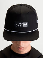 Alpinestars Label Snapback, casquette de sportswear lifestyle unisexe, noire et blanche, en sergé de coton, avec couronne structurée, visière plate, cordon blanc et écusson tissé Alpinestars, portée par un modèle