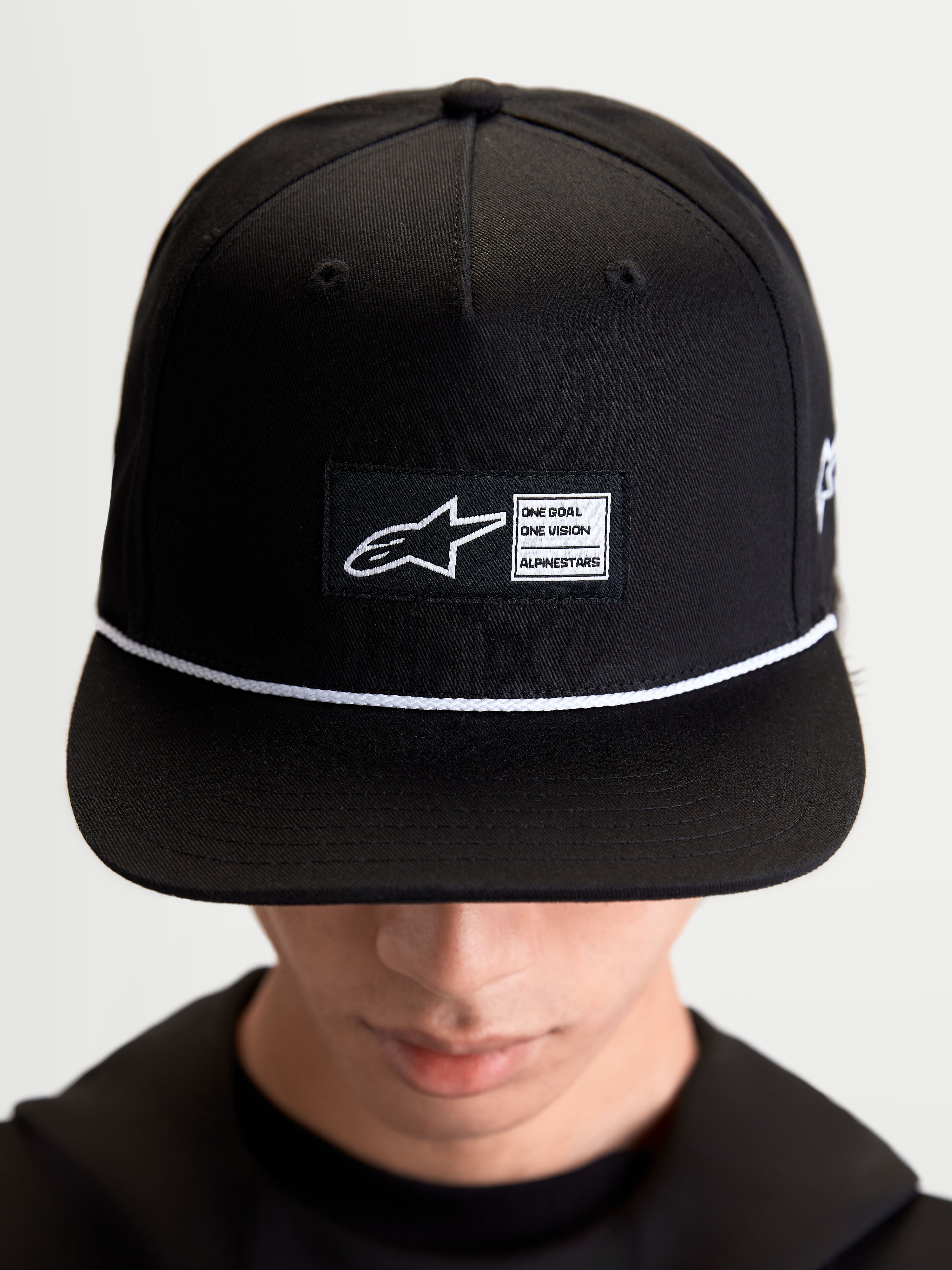 Alpinestars Label Snapback, casquette de sportswear lifestyle unisexe, noire et blanche, en sergé de coton, avec couronne structurée, visière plate, cordon blanc et écusson tissé Alpinestars, portée par un modèle