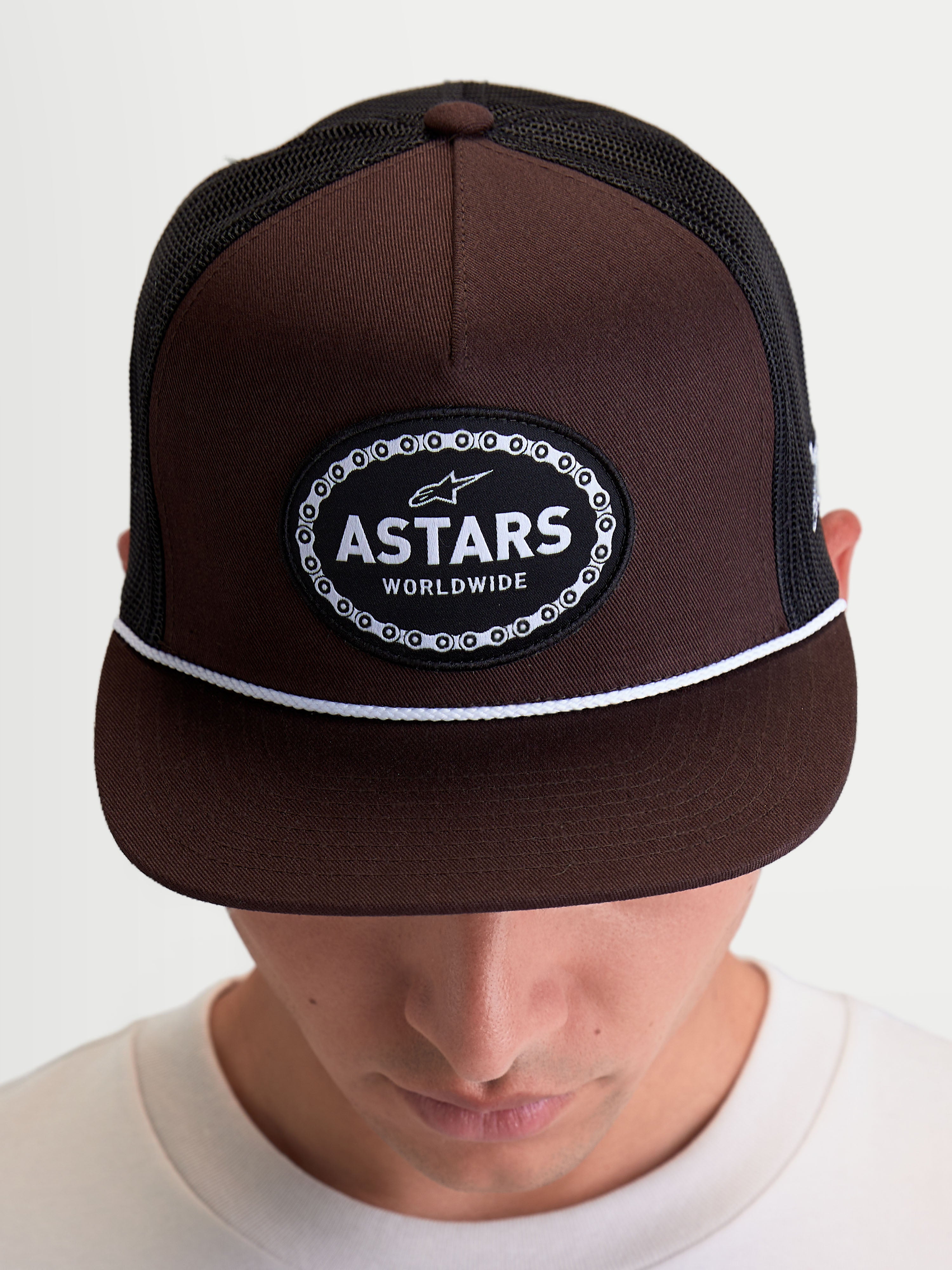 Alpinestars Hotlink Trucker pet, motorsport-geïnspireerde sportswear-pet getoond op model, bruin en zwart, voorzien van een gestructureerde kroon, platte klep met wit koorddetail en een Astars Worldwide-logopatch met fietskettingrand.