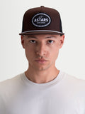 Alpinestars Hotlink trucker pet, bruin en zwarte motorsportpet, met een gestructureerde kroon, platte klep met wit touwdetail en een geweven Astars Worldwide logo patch op de voorkant, gedragen door een model.