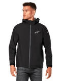 Männliches Model trägt Alpinestars Kascade Regenjacke, Schwarz, weißes Brustlogo und Ärmel-Branding, Regenjacke mit Kapuze, beschichtetem Frontreißverschluss und Klettverschlussbündchen, kombiniert mit dunkler Jeans, lässige Motorsport-inspirierte Streetwear