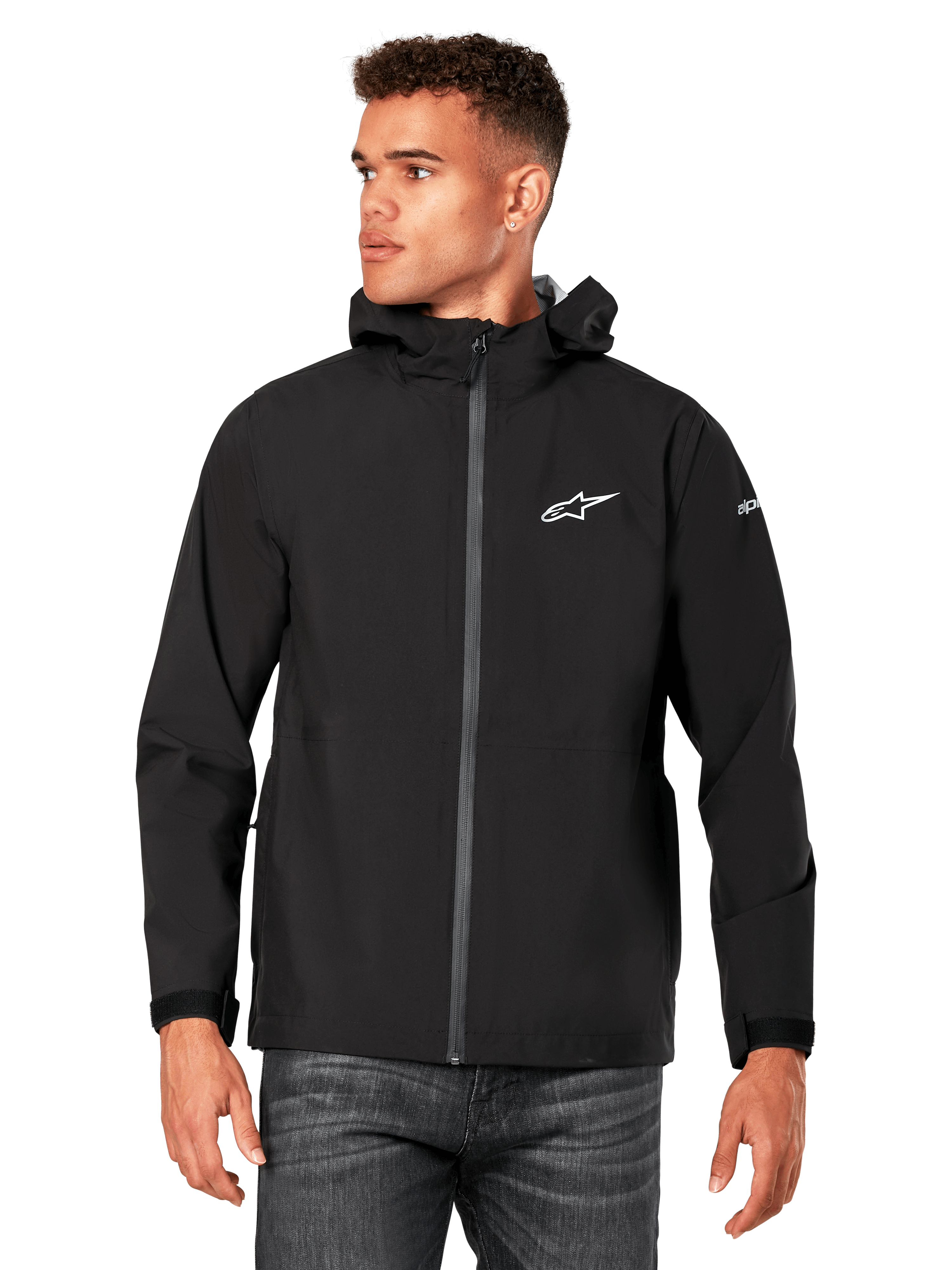 Männliches Model trägt Alpinestars Kascade Regenjacke, Schwarz, weißes Brustlogo und Ärmel-Branding, Regenjacke mit Kapuze, beschichtetem Frontreißverschluss und Klettverschlussbündchen, kombiniert mit dunkler Jeans, lässige Motorsport-inspirierte Streetwear