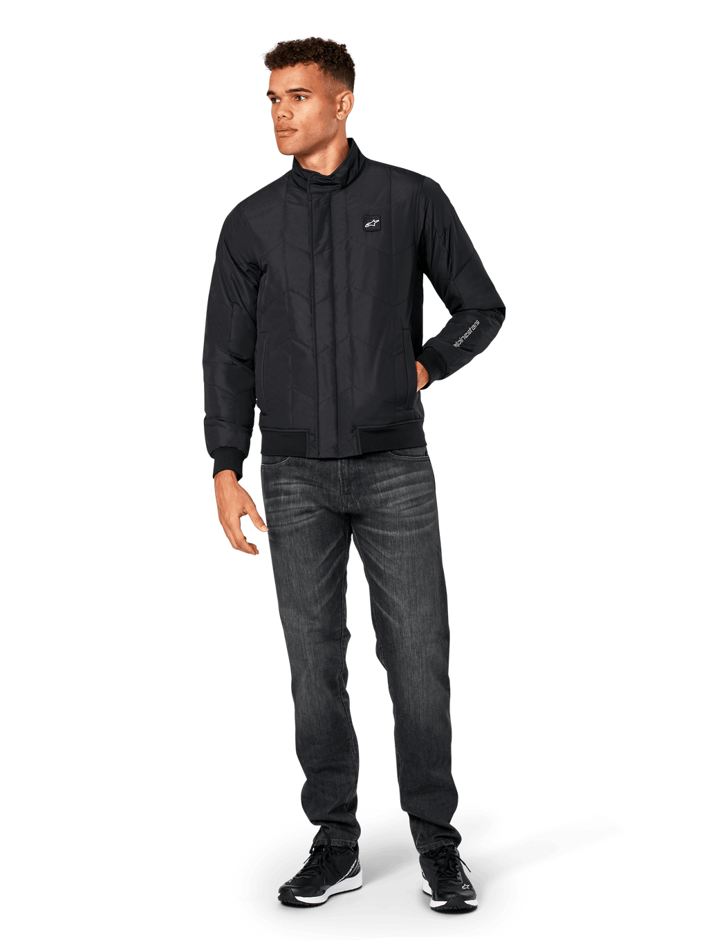 Alpinestars Precedent jas, gesteppte sportswear jas, zwart, gedragen met donkergrijze jeans en zwarte sneakers, voorzien van een schuin gestikt patroon, lage kraag, ribgebreide manchetten en zoom, met een witte logopatch op de borst en borduurwerk op de mouw.