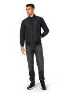 Alpinestars Precedent jas, gesteppte sportswear jas, zwart, gedragen met donkergrijze jeans en zwarte sneakers, voorzien van een schuin gestikt patroon, lage kraag, ribgebreide manchetten en zoom, met een witte logopatch op de borst en borduurwerk op de mouw.